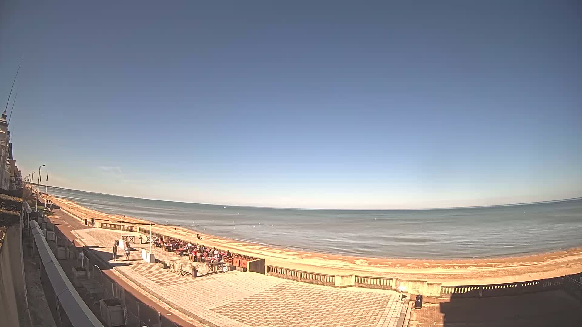 A sunny day shows a beachfront patio with people sitting at outdoor tables, overlooking a calm ocean and sandy beach.