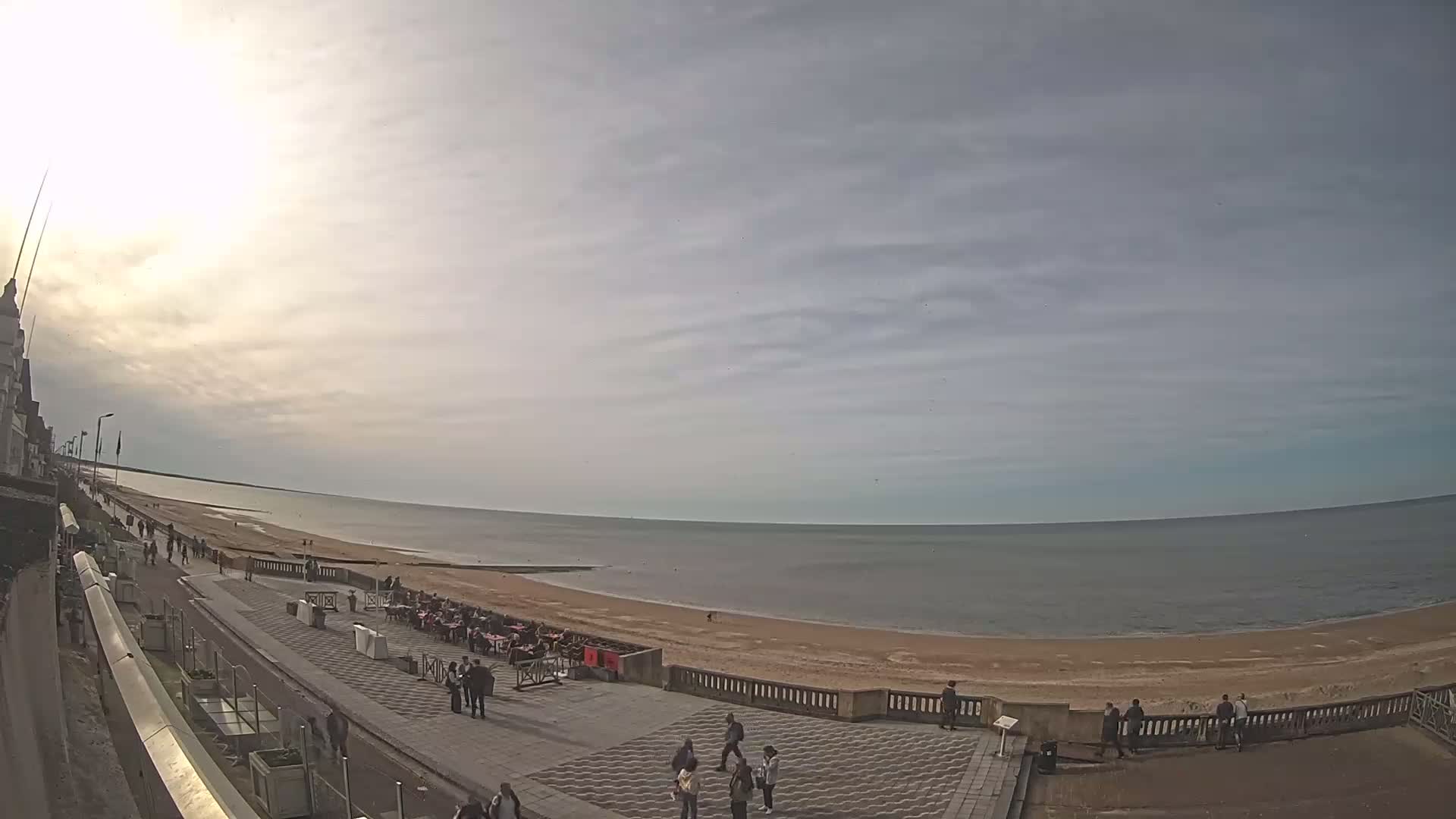 A mostly sunny day at a seaside promenade shows people walking along a paved walkway next to a sandy beach and calm ocean.