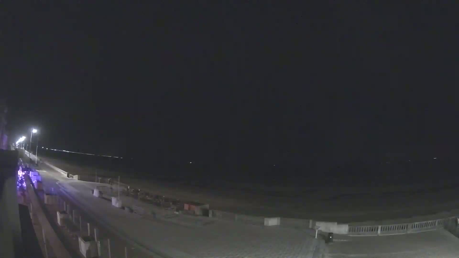 A nighttime view of a mostly empty beach and boardwalk, illuminated by streetlights, under a clear, dark sky.