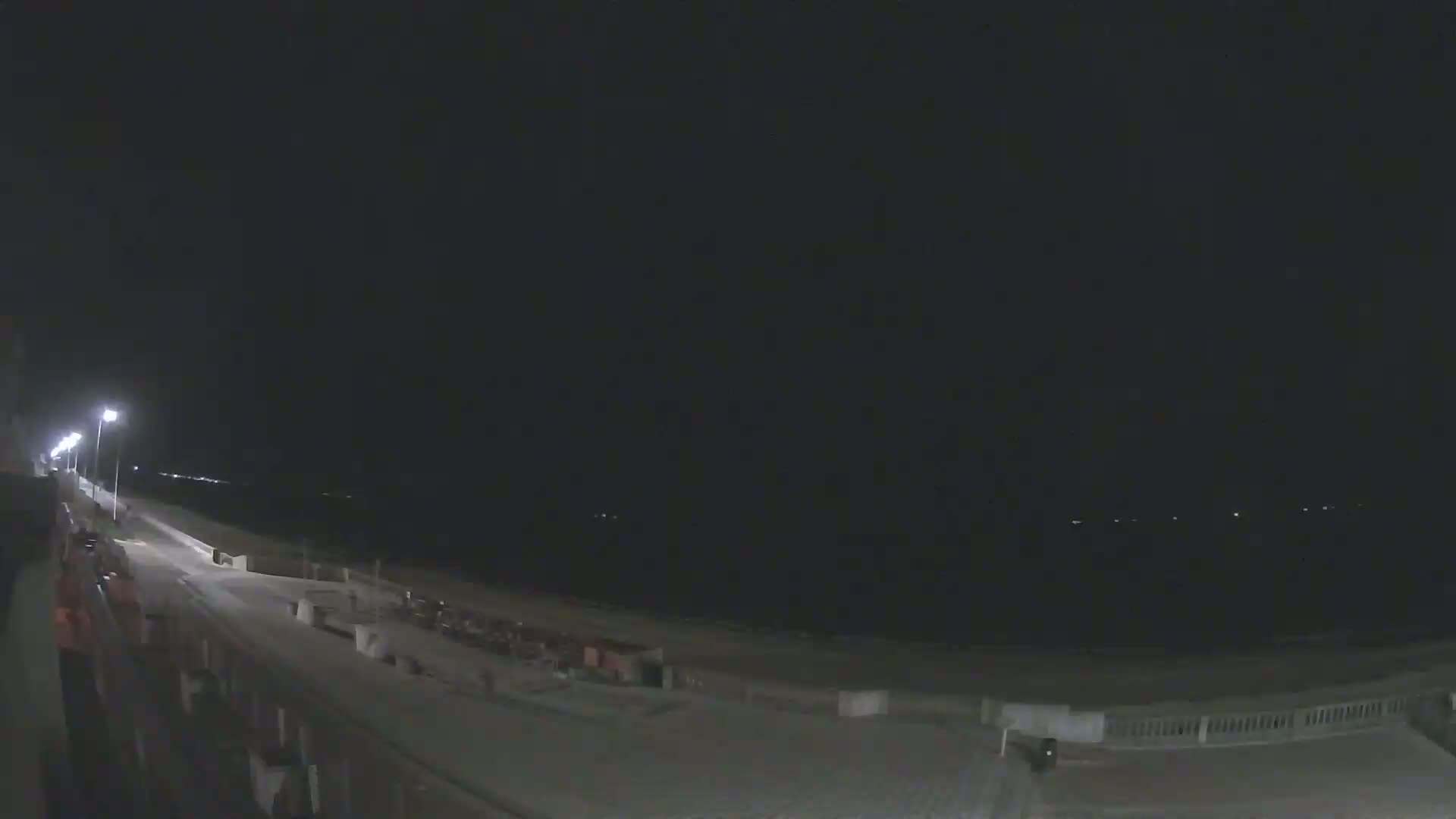 A nighttime view of a mostly empty boardwalk and beach, lit by streetlights, under a dark, clear sky.