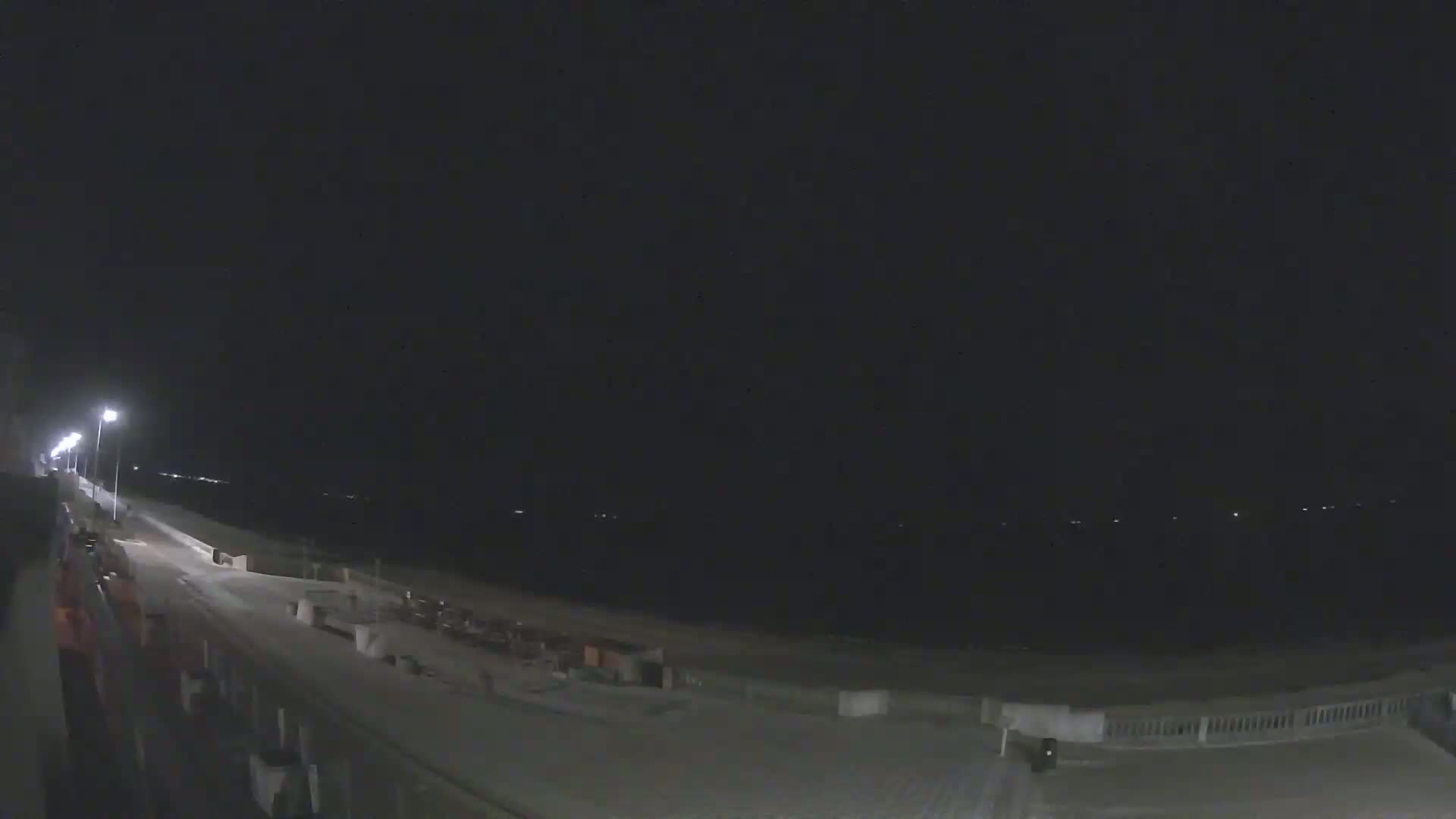 A nighttime view of a mostly empty boardwalk and beach, illuminated by streetlights.