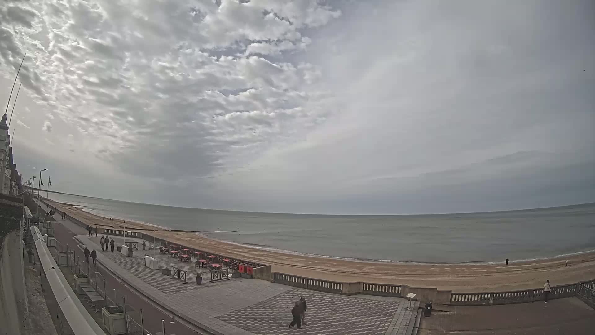 A mostly cloudy day shows a wide, sandy beach with a calm sea, a paved walkway with a few people walking, and outdoor seating at restaurants near the water.