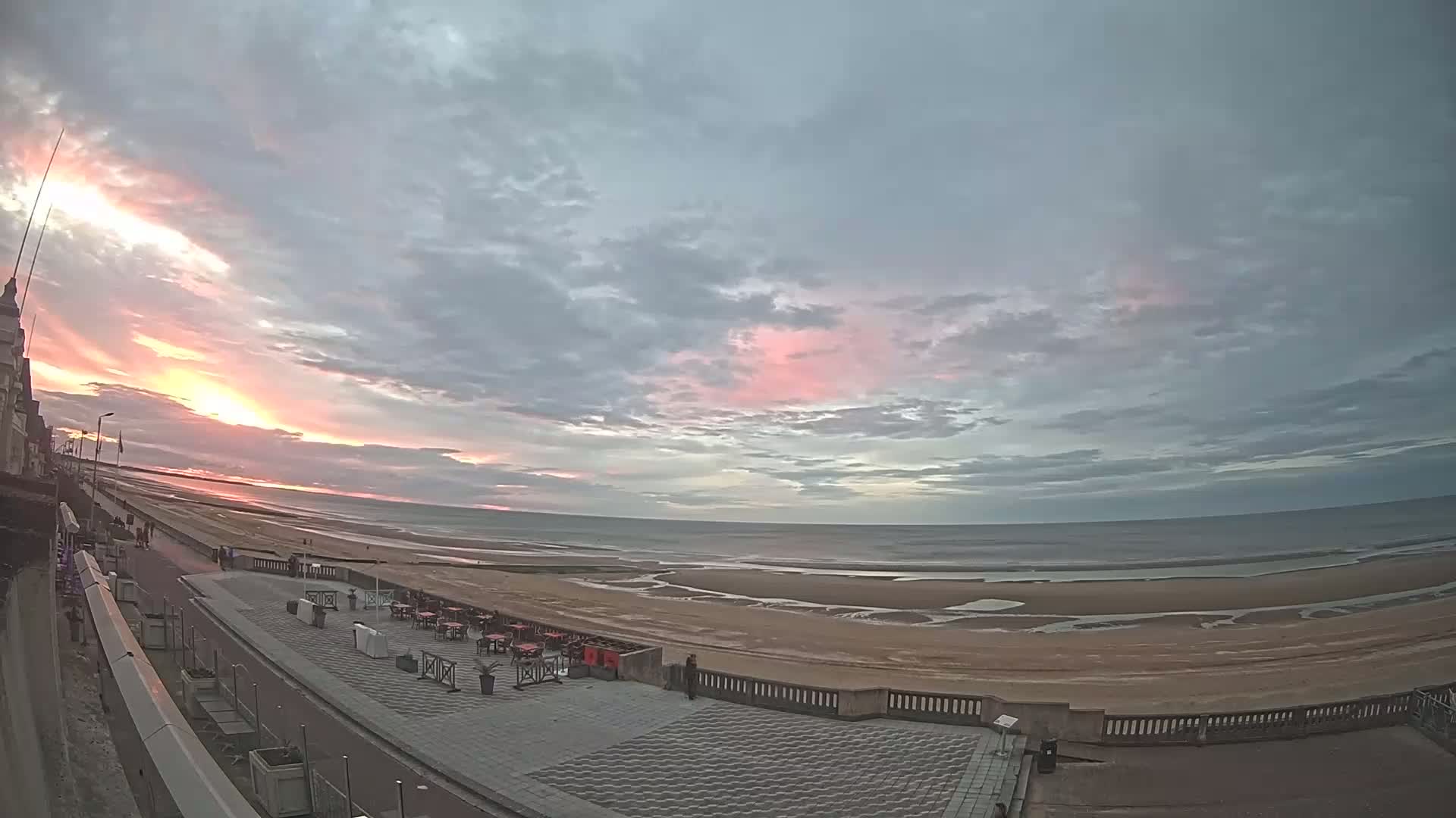 A beach with a promenade and outdoor seating area is visible at sunset under a partly cloudy sky.