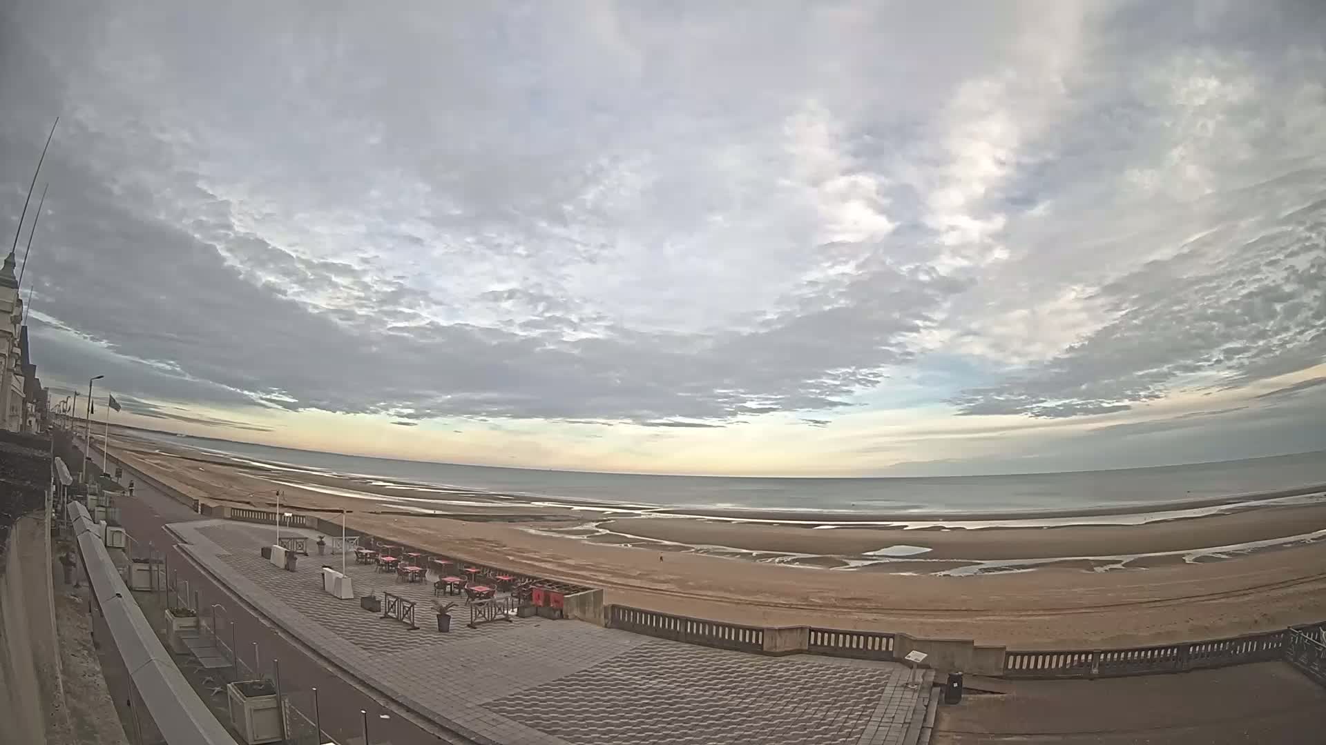 A wide, overcast sky over a sandy beach with a promenade lined with outdoor seating, showing a calm ocean in the distance.