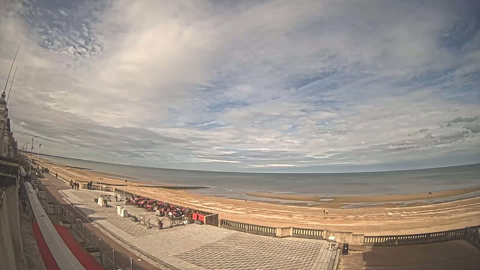 A mostly sunny day at a beachside promenade shows a wide sandy beach, calm ocean, and outdoor seating area with several people present.