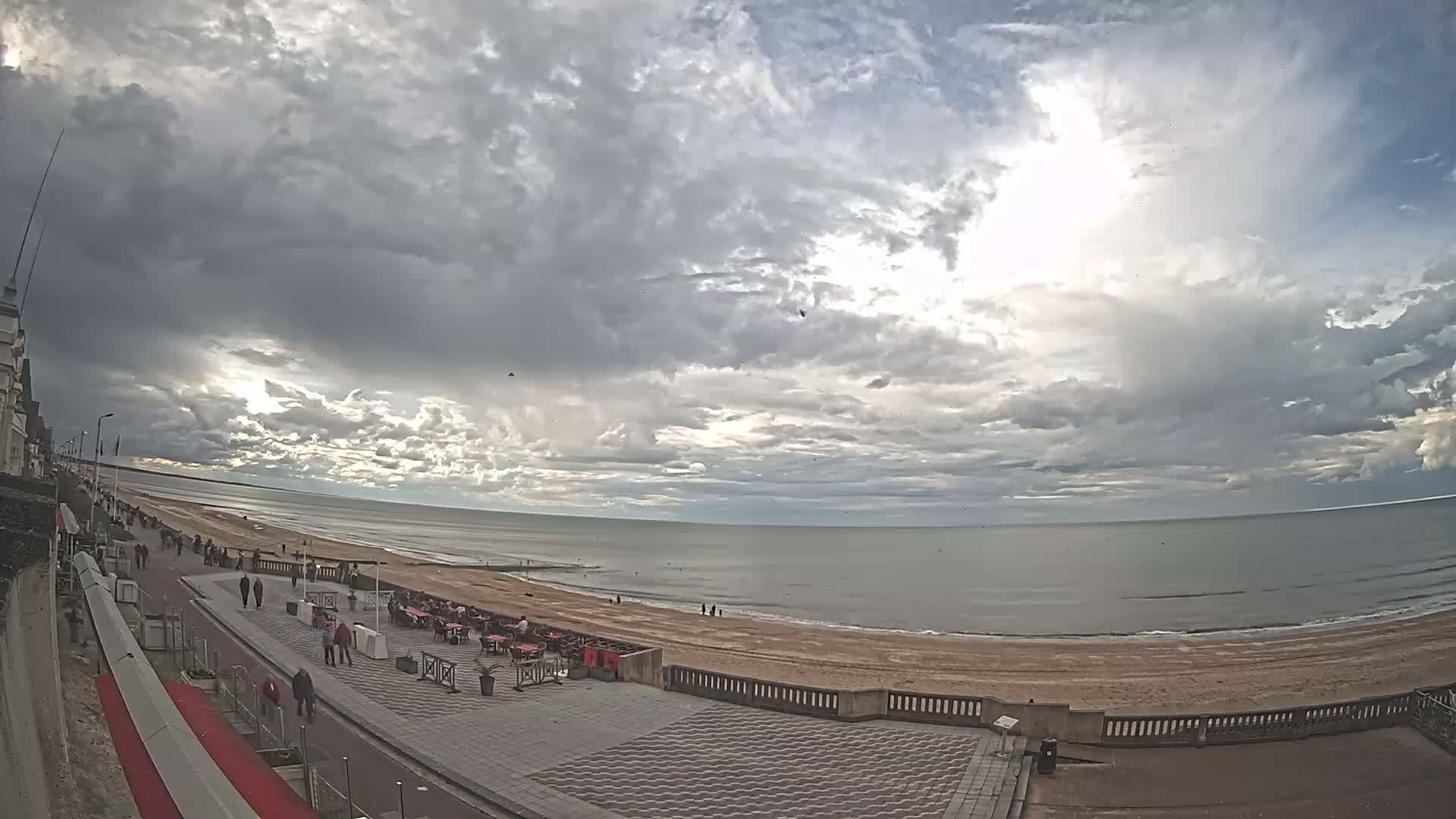 A mostly cloudy sky overlooks a calm ocean beach with people walking along a paved boardwalk next to outdoor seating areas.