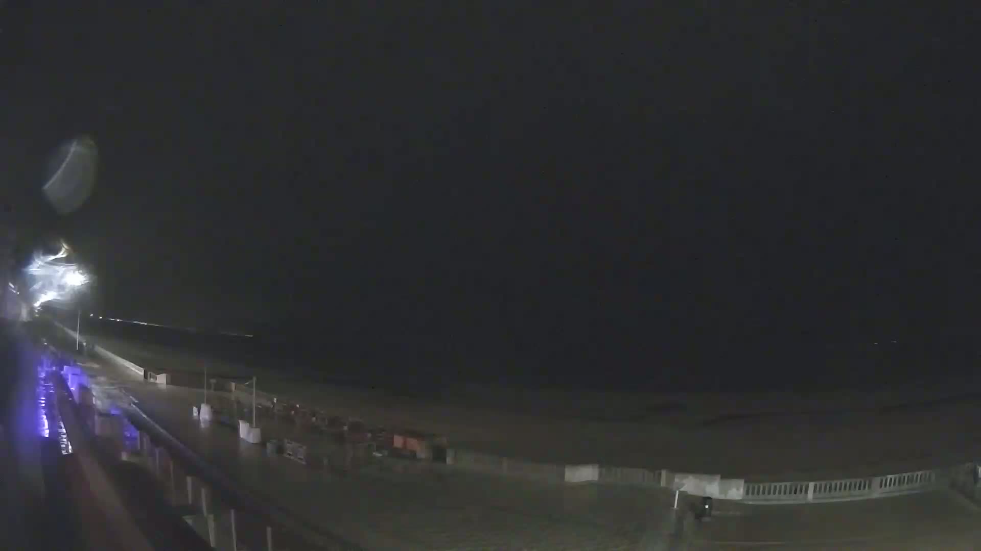 A nighttime view of a wet beach with a boardwalk and some structures, under a dark sky.