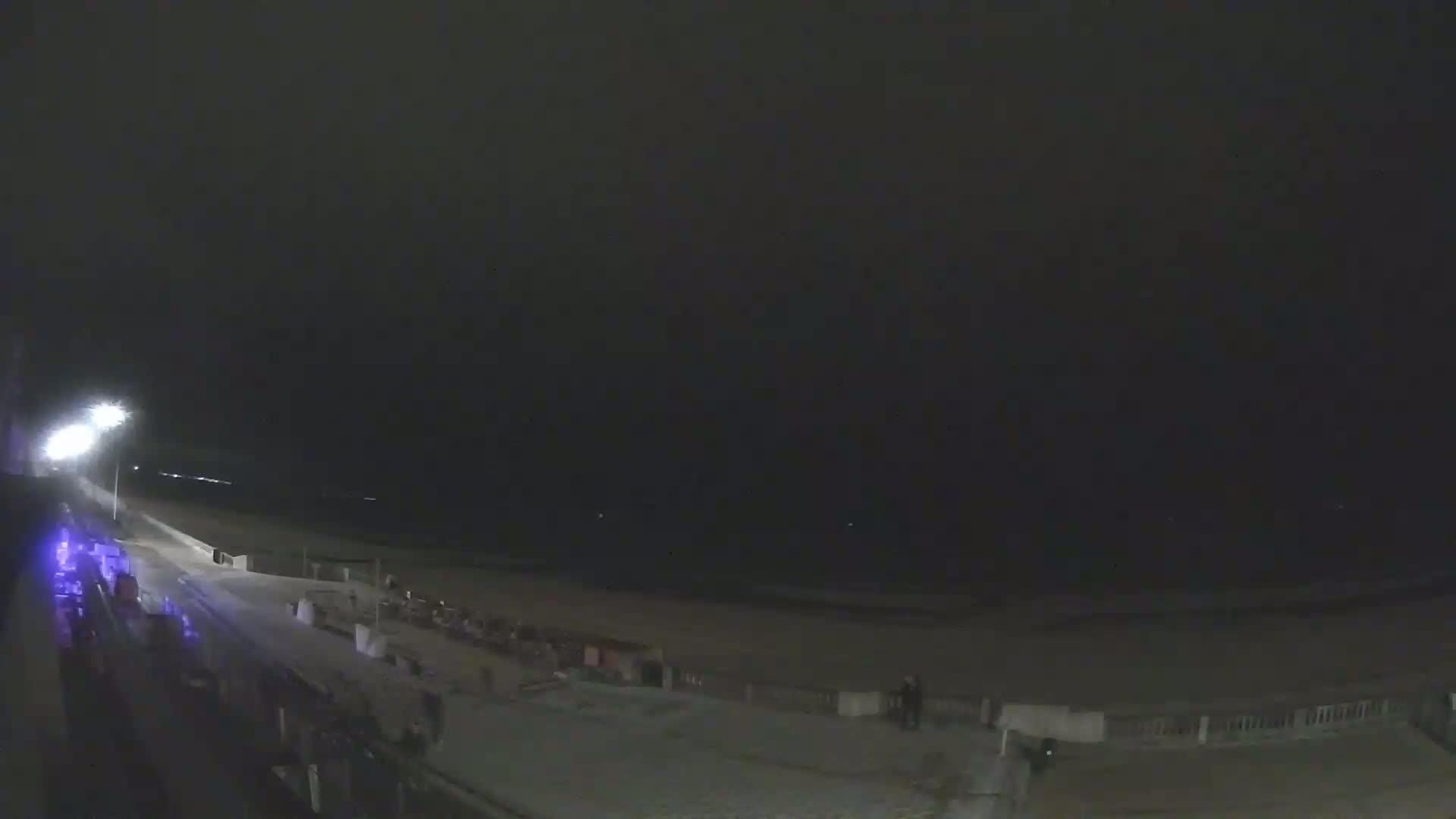 A dark, nighttime scene shows a beach with a paved walkway, sparsely populated with a few people, under a cloudy sky.