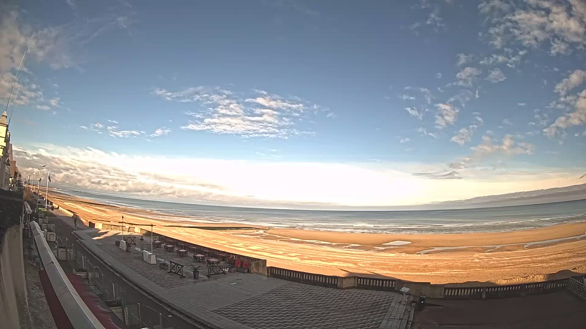 A mostly sunny day shows a wide sandy beach with a calm ocean, viewed from a paved boardwalk lined with outdoor seating.
