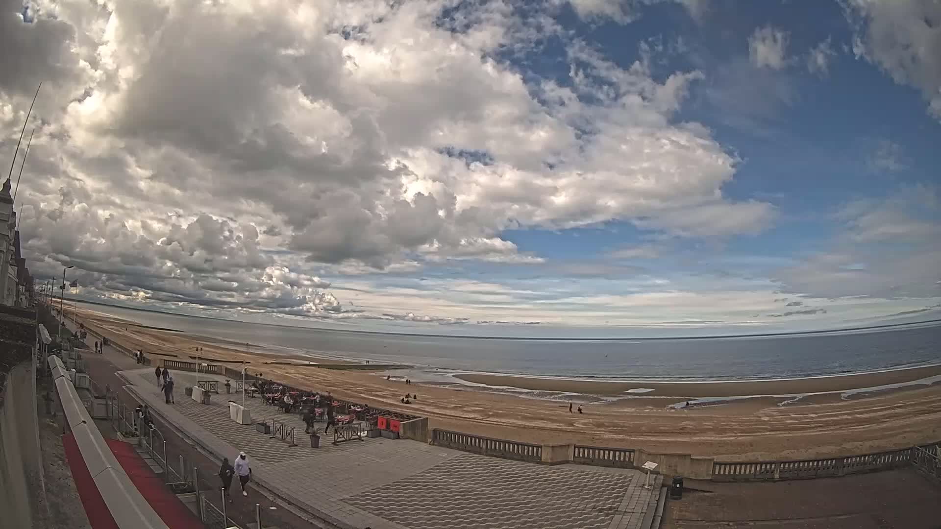 A partly cloudy sky overlooks a wide sandy beach and calm ocean, with people strolling along a paved promenade lined with outdoor seating.