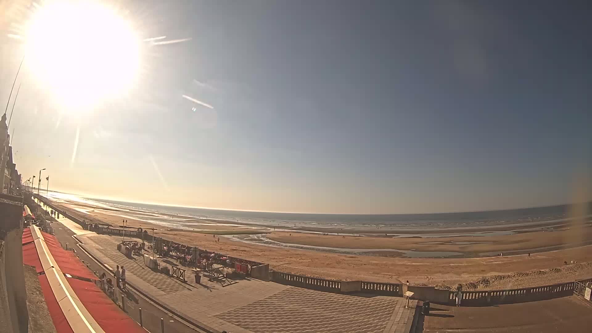 A sunny day at a beach, showing a wide sandy shore, calm ocean waters, and people strolling along a paved beachfront promenade with outdoor seating areas.