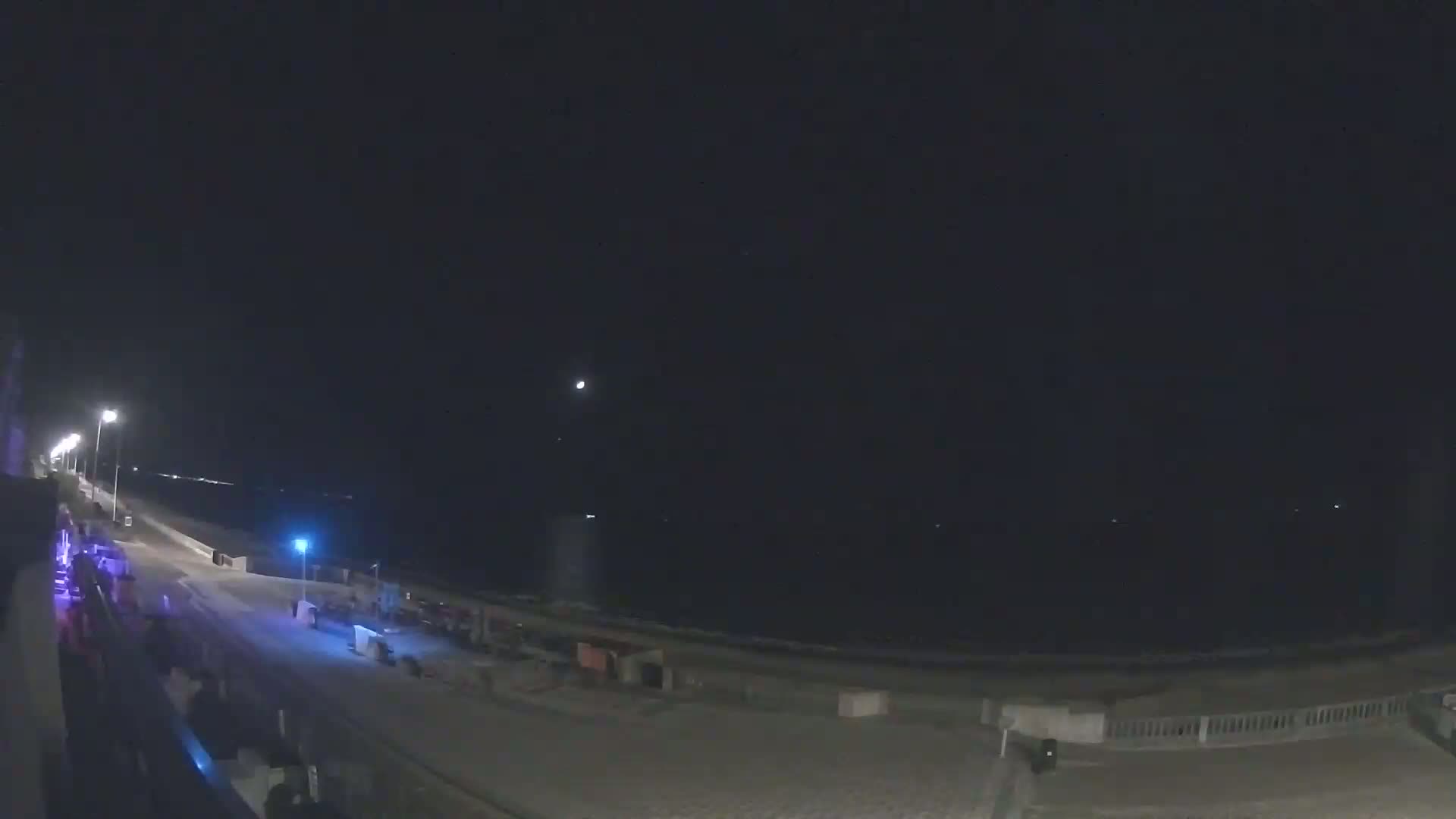 A nighttime view of a beach with a few lights along a boardwalk, under a mostly clear, dark sky.