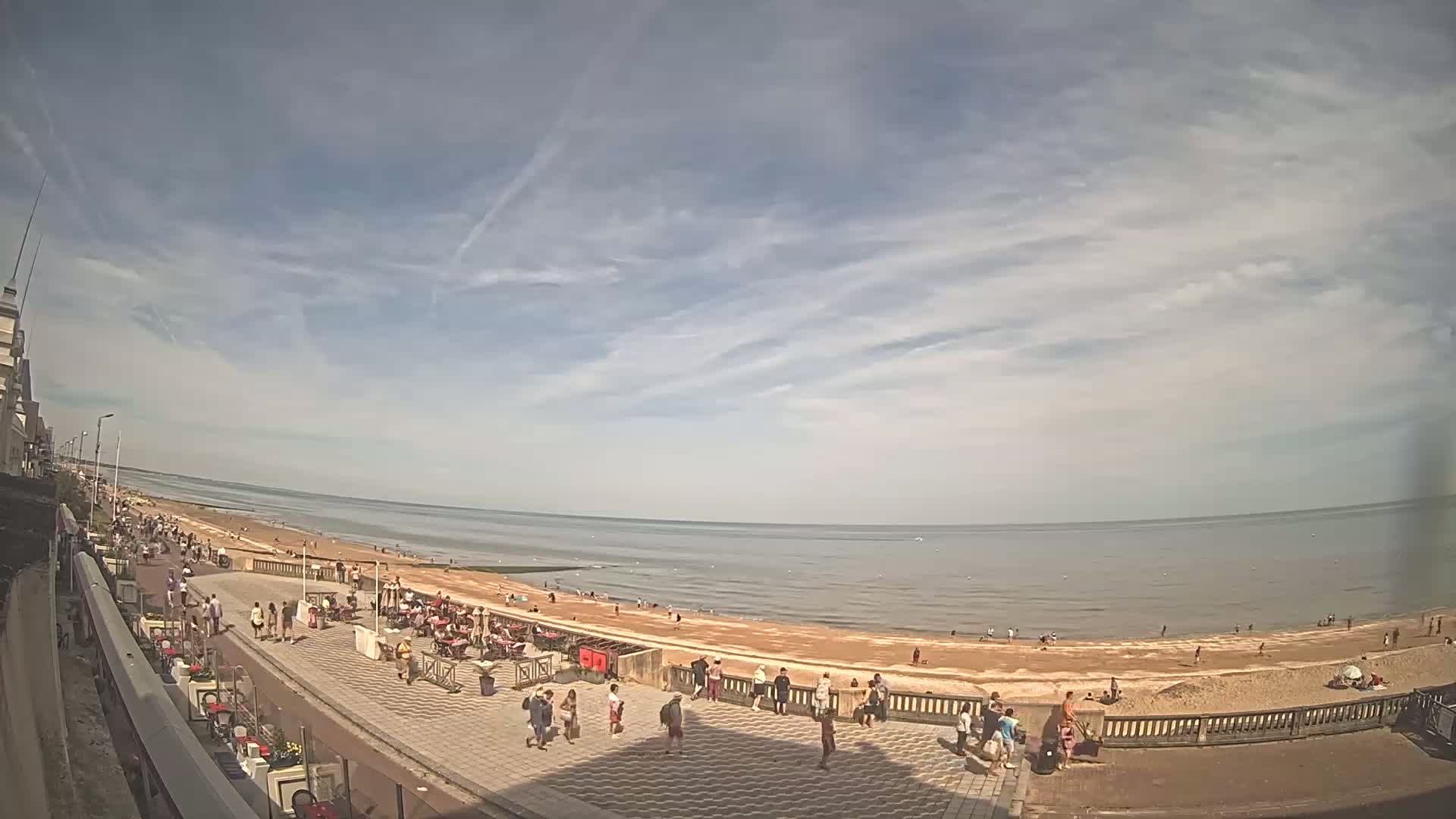 A partly cloudy day at a beach with many people on the sand and a paved walkway lined with outdoor seating.