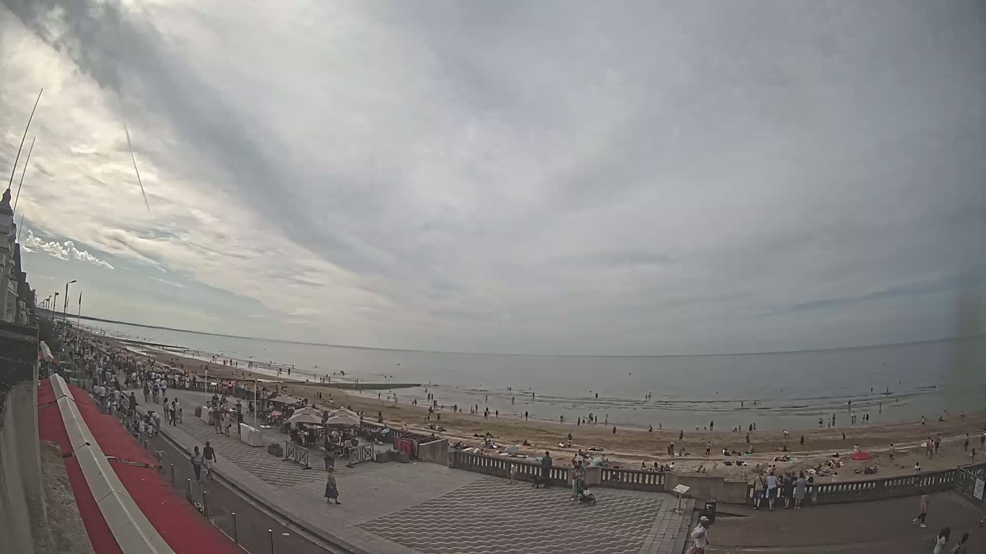 A mostly cloudy day at a beach shows many people on the sand and walking along the boardwalk near the water.