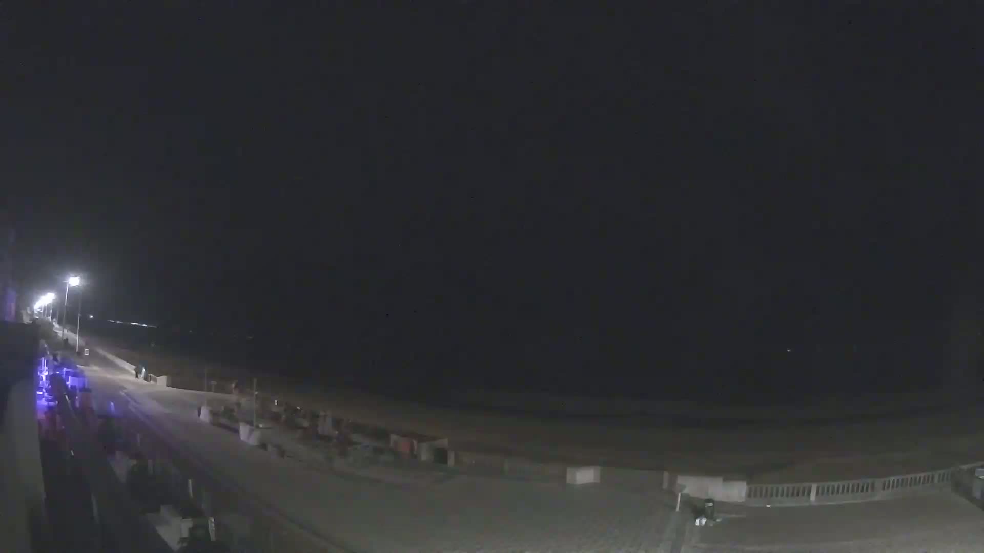 A nighttime view of a mostly empty boardwalk and beach, illuminated by streetlights.