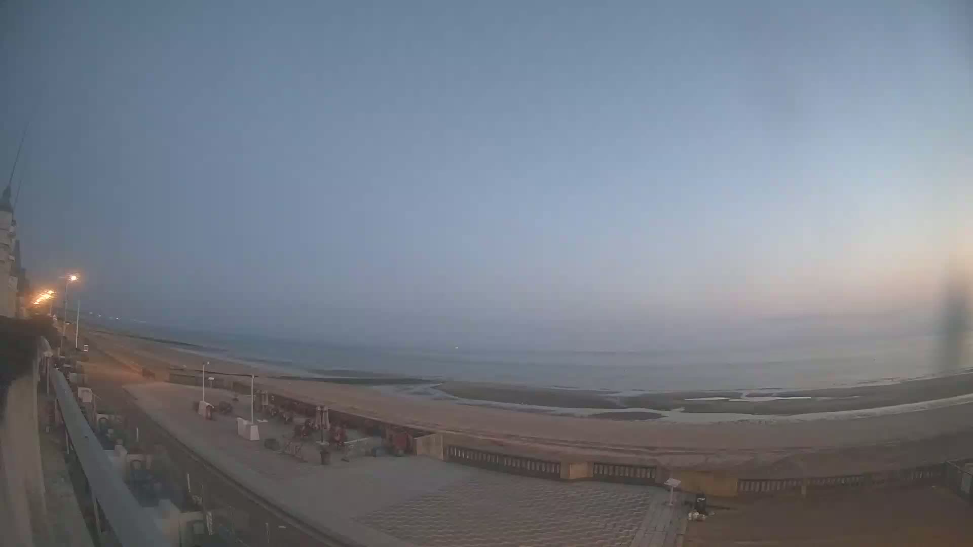 A hazy twilight scene shows a beach with a paved walkway and some buildings, overlooking a calm ocean.