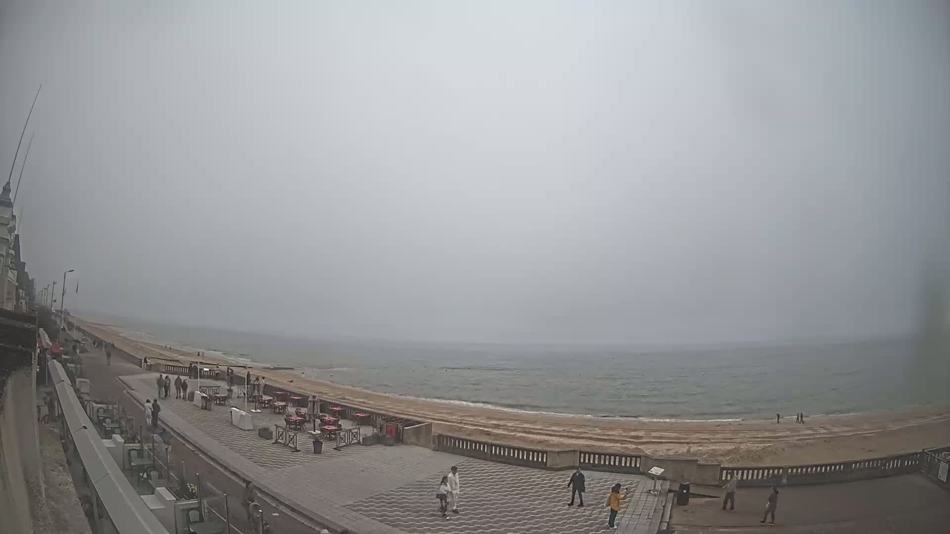 A hazy, overcast day shows a beach with a paved promenade, outdoor seating, and a few people walking along the shore.