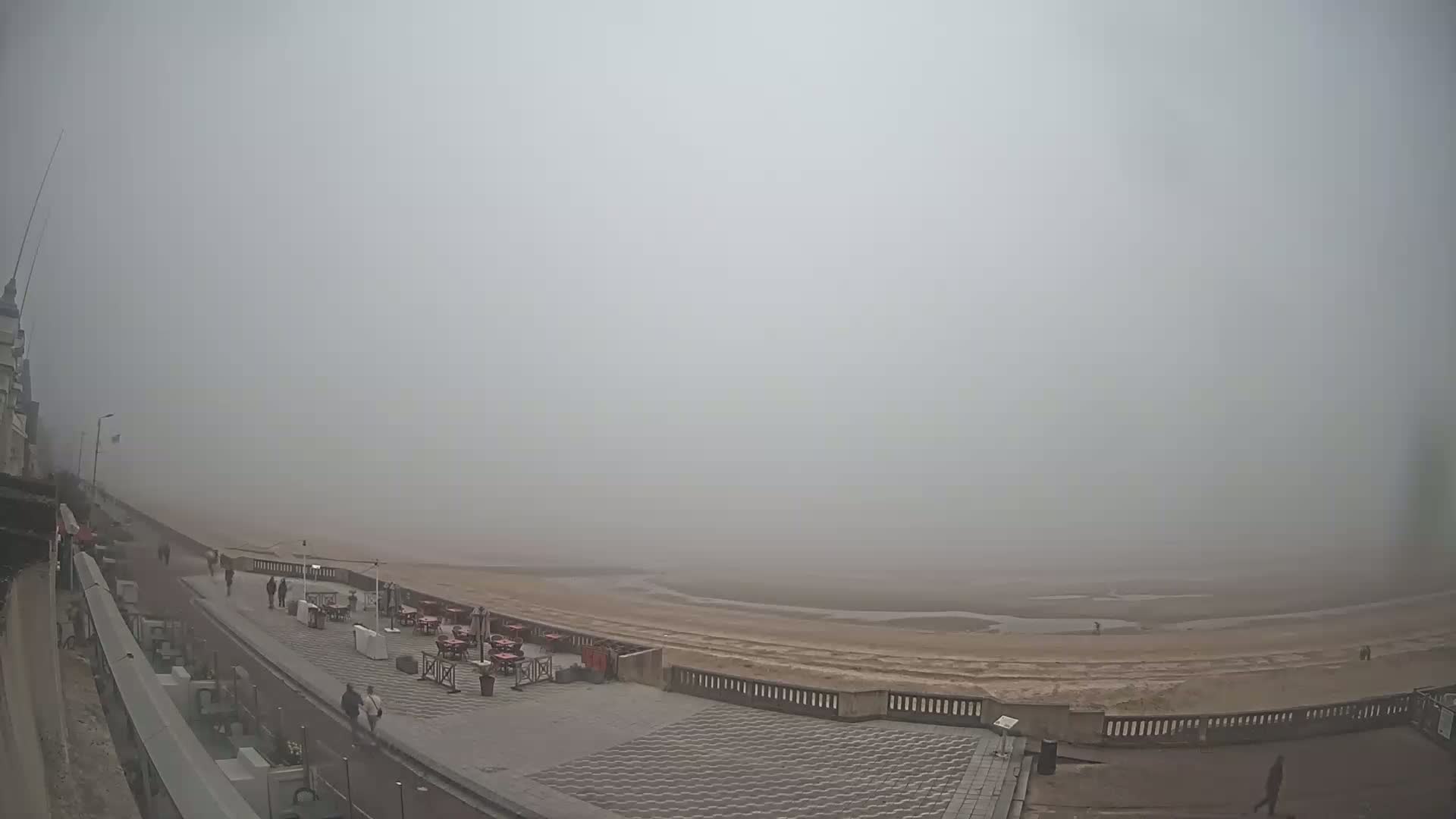 A foggy beach scene shows a paved walkway next to a sandy beach with several outdoor tables and chairs, and a few people walking along the path.