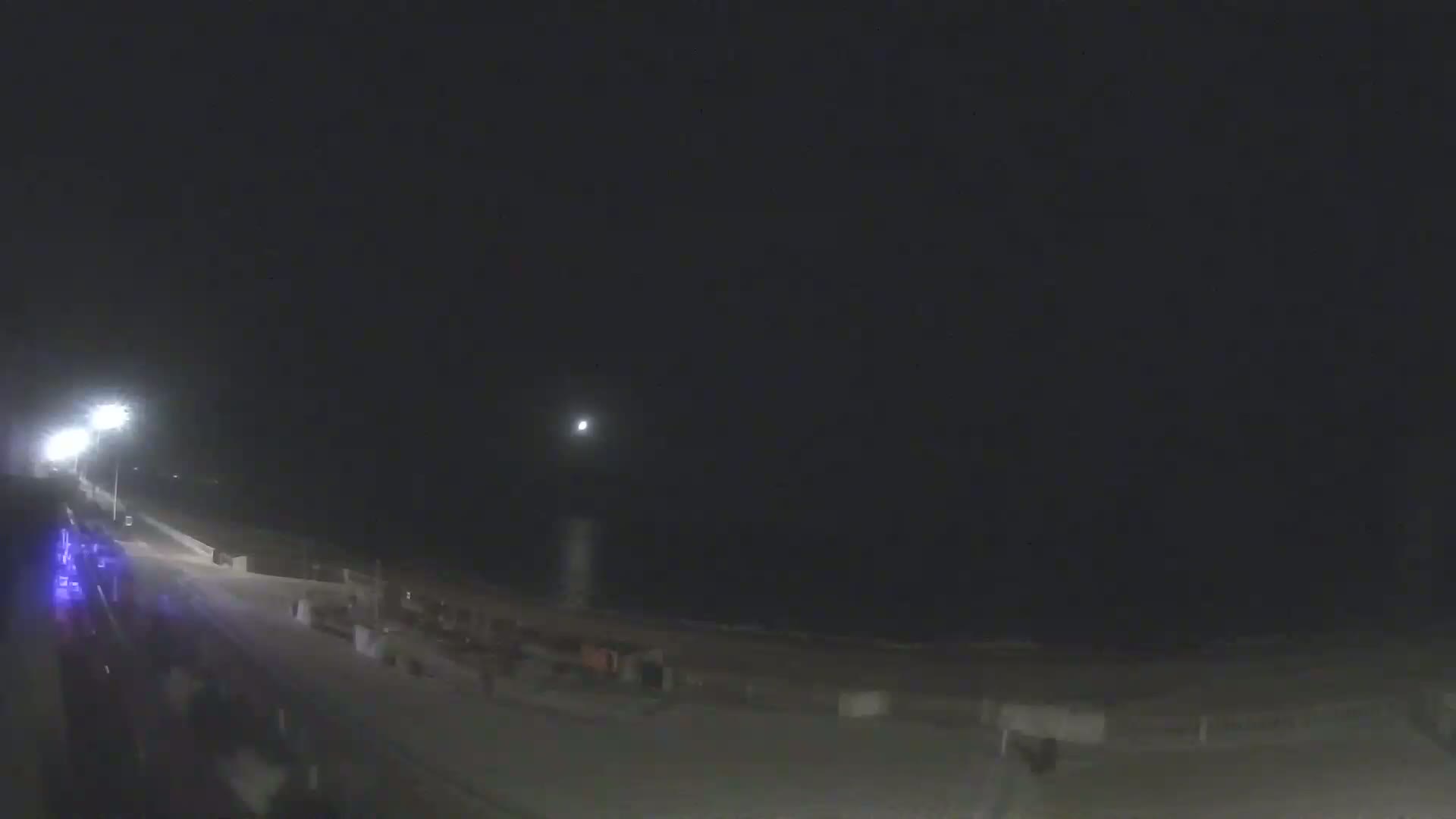 A nighttime view of a beach with a road and some structures visible under a dark, moonlit sky.