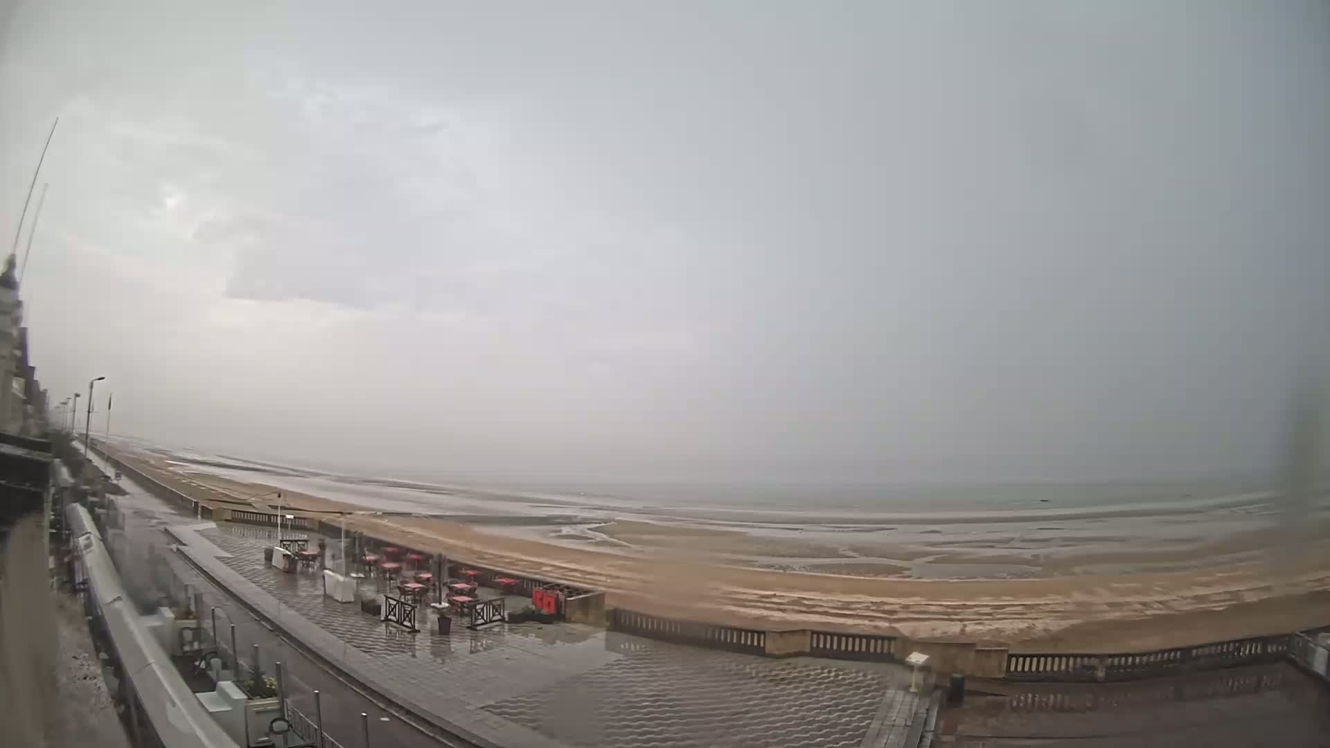 A wet, paved promenade overlooks a wide, sandy beach and calm sea under an overcast sky.