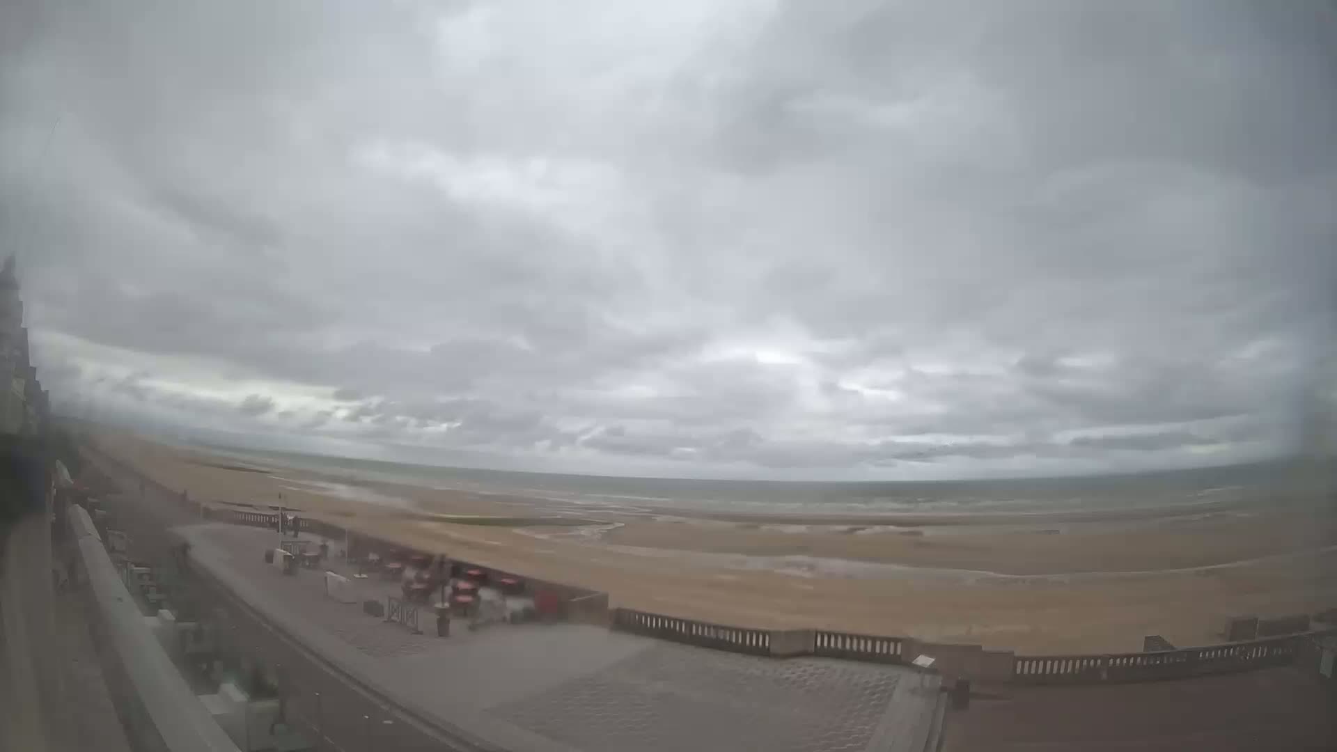 A wide shot shows an overcast sky over a beach with a paved walkway and outdoor seating areas, under what appears to be a light rain or drizzle.