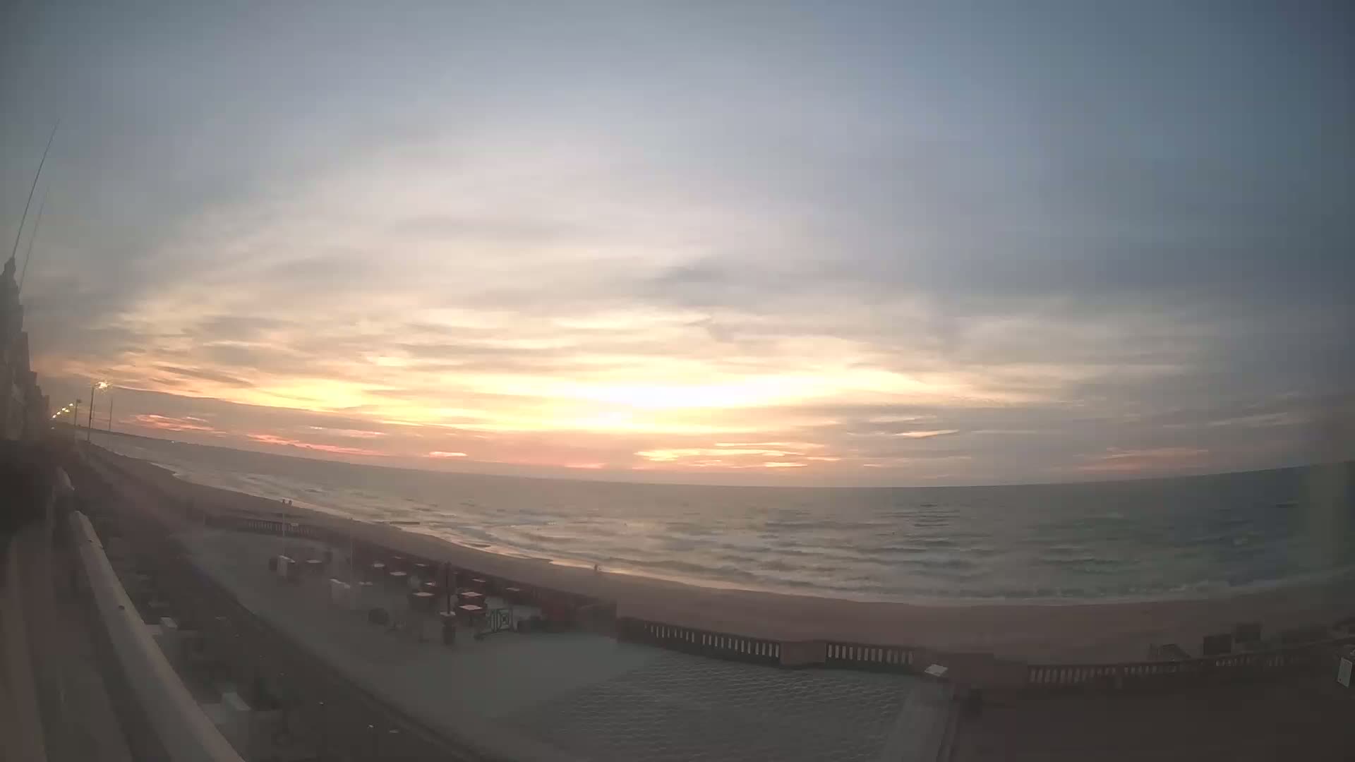 A sunrise over a calm ocean beach with a boardwalk and some outdoor furniture visible.