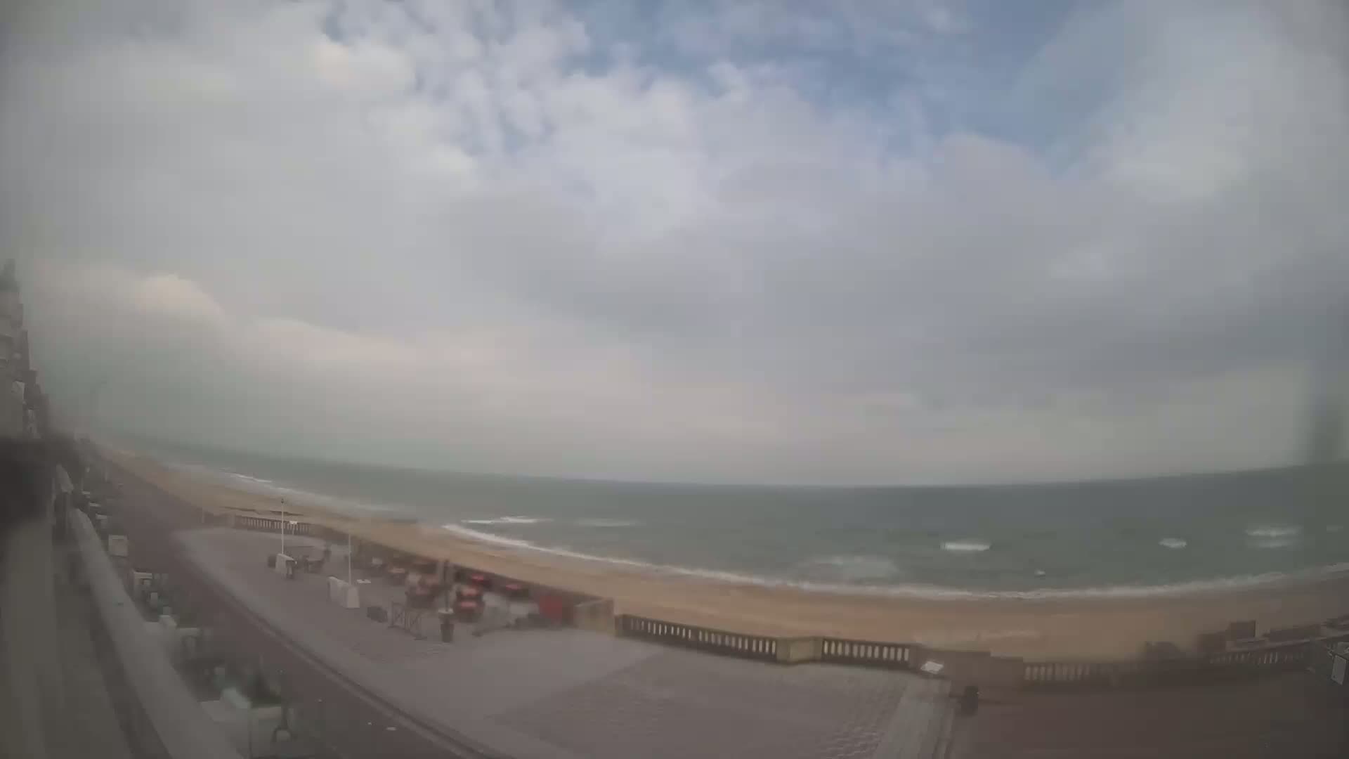 A mostly cloudy day shows a beach with a calm sea, viewed from a high vantage point overlooking a paved walkway.