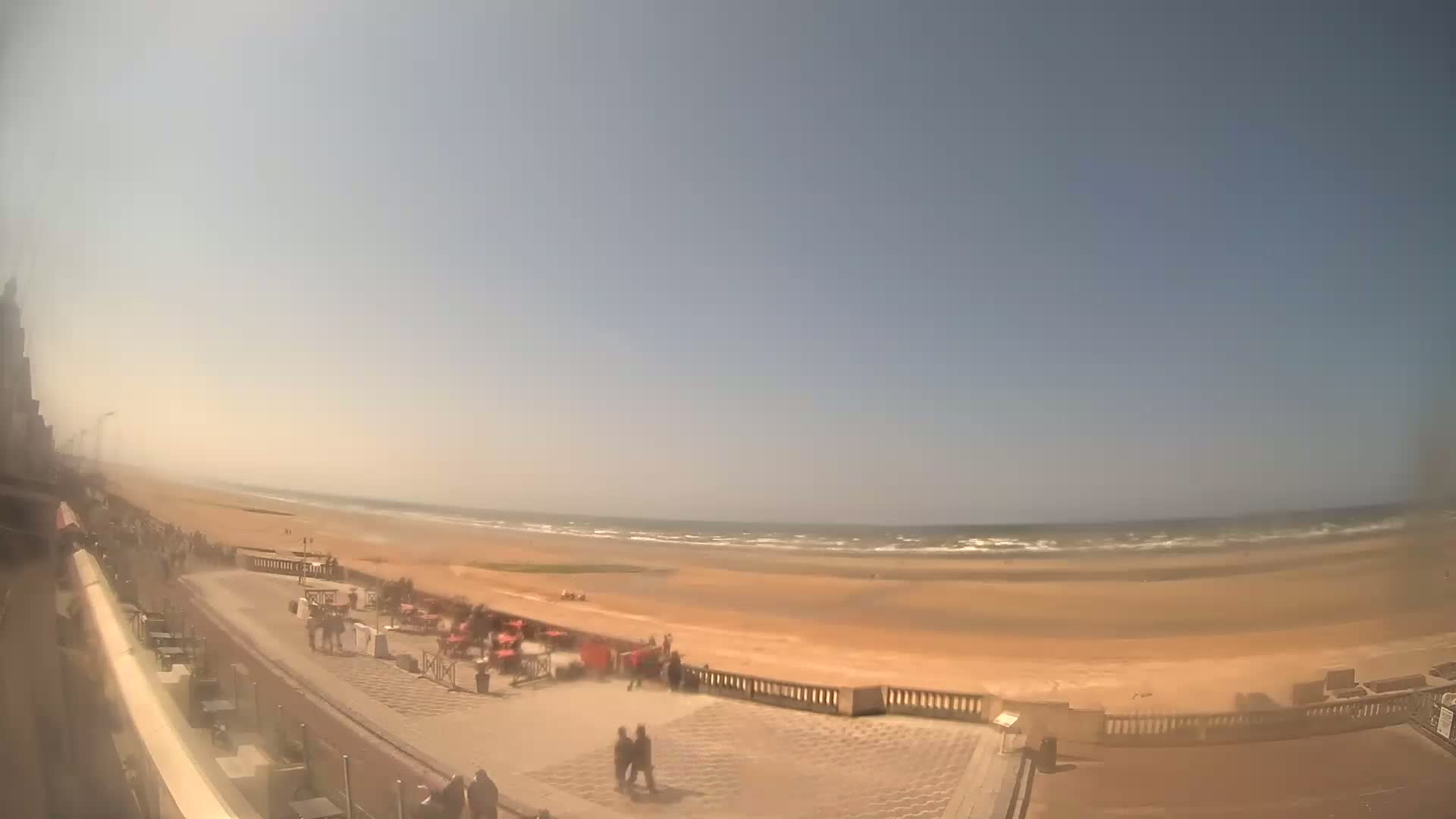 A hazy, sunny day at a beach with a wide sandy shore, gentle waves, and people strolling along a paved promenade near some outdoor seating.