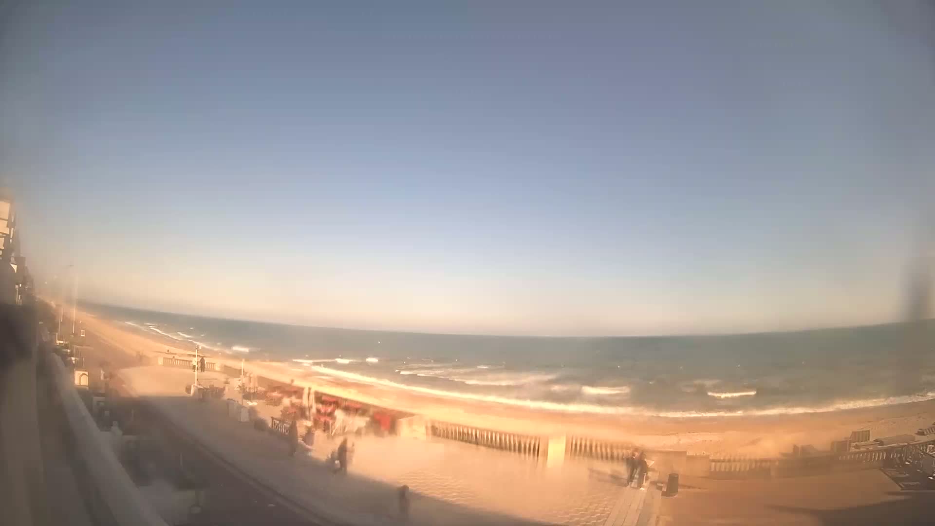 A mostly clear, sunny day at a beach with moderate waves, showing a boardwalk with a few people walking along it.