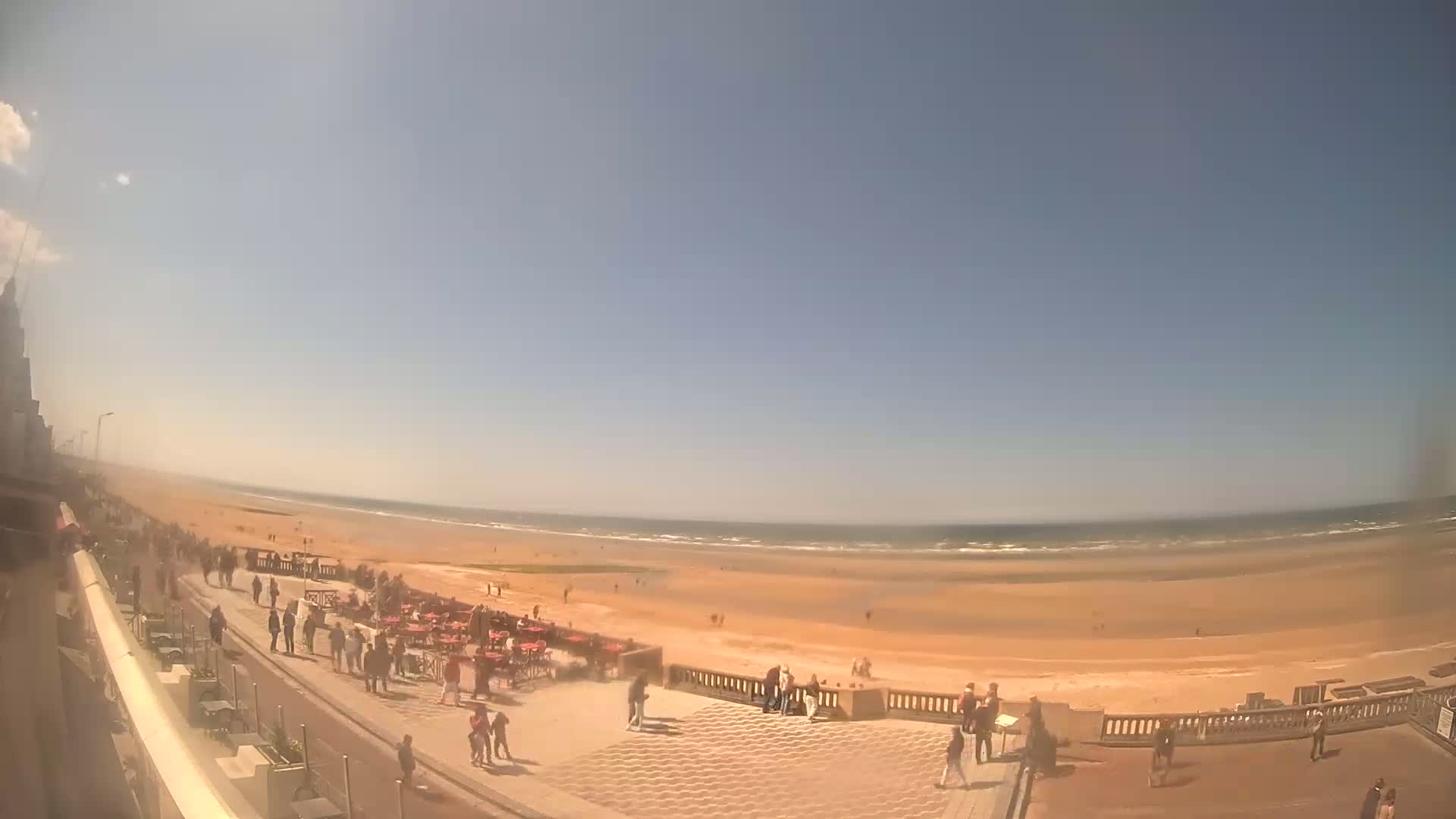 A sunny day at a beach shows people walking along a boardwalk near the ocean, with some people seated at outdoor tables.