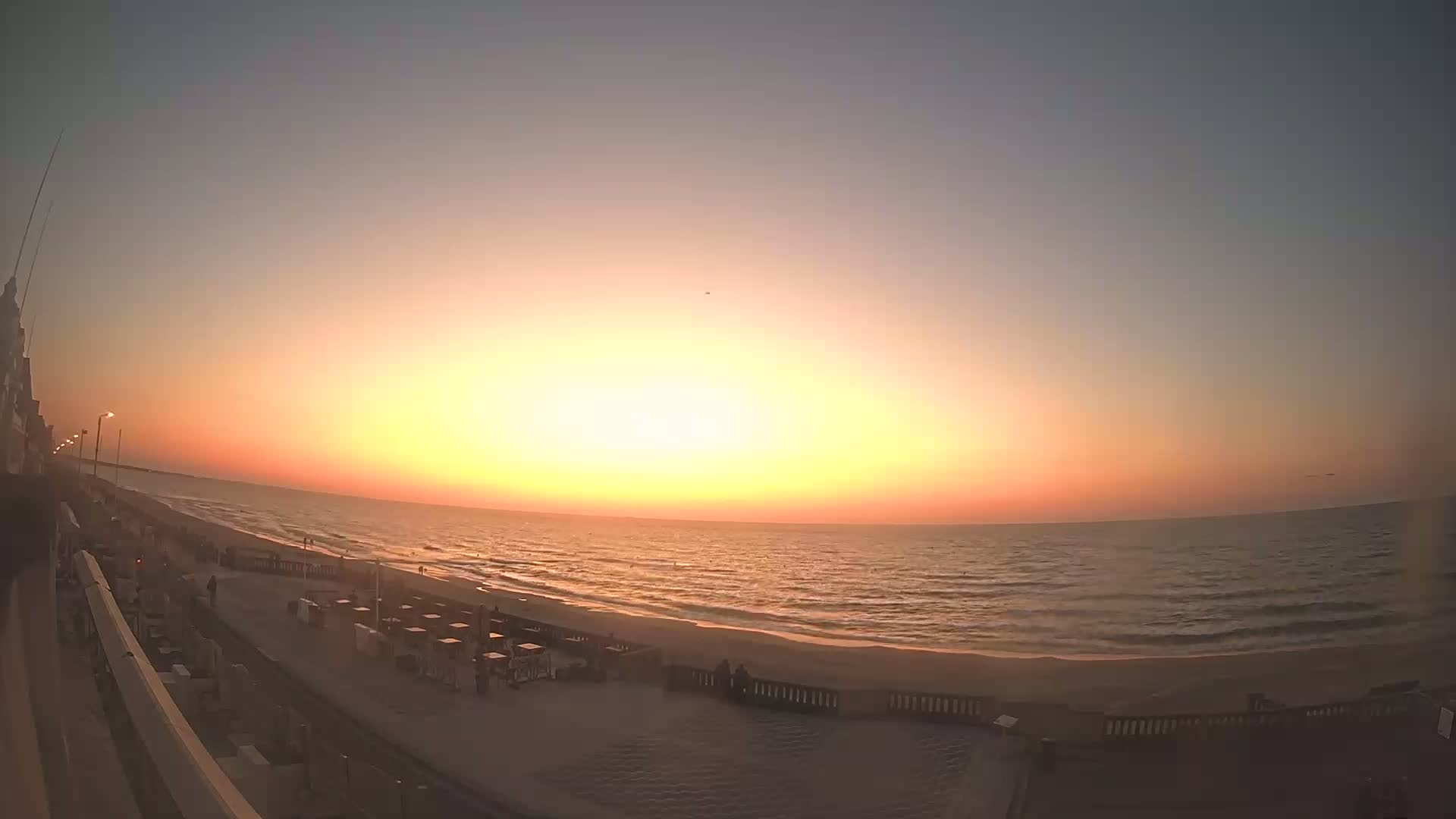 A wide shot of an ocean beach at sunset, with a boardwalk and several people visible.