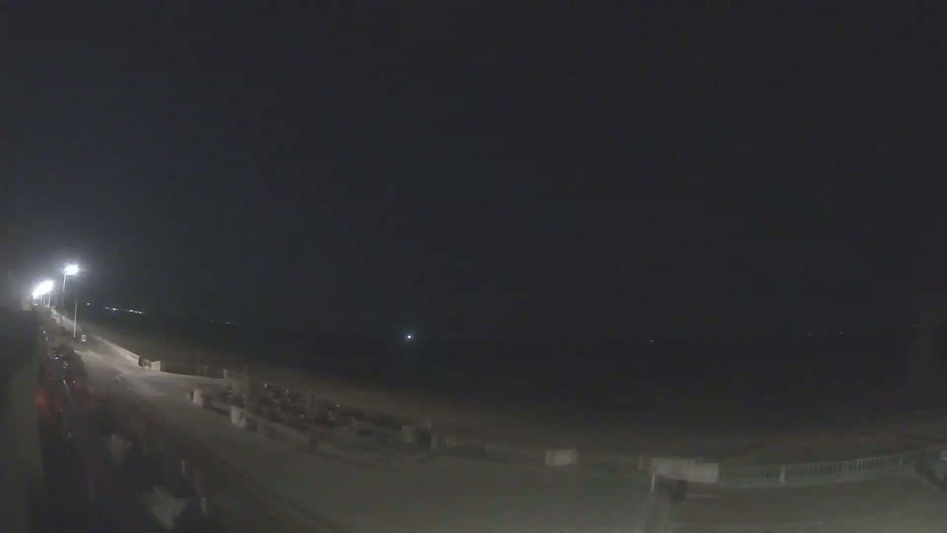 A nighttime view of a beach at night, showing a mostly empty roadway and a dark ocean under a clear sky.