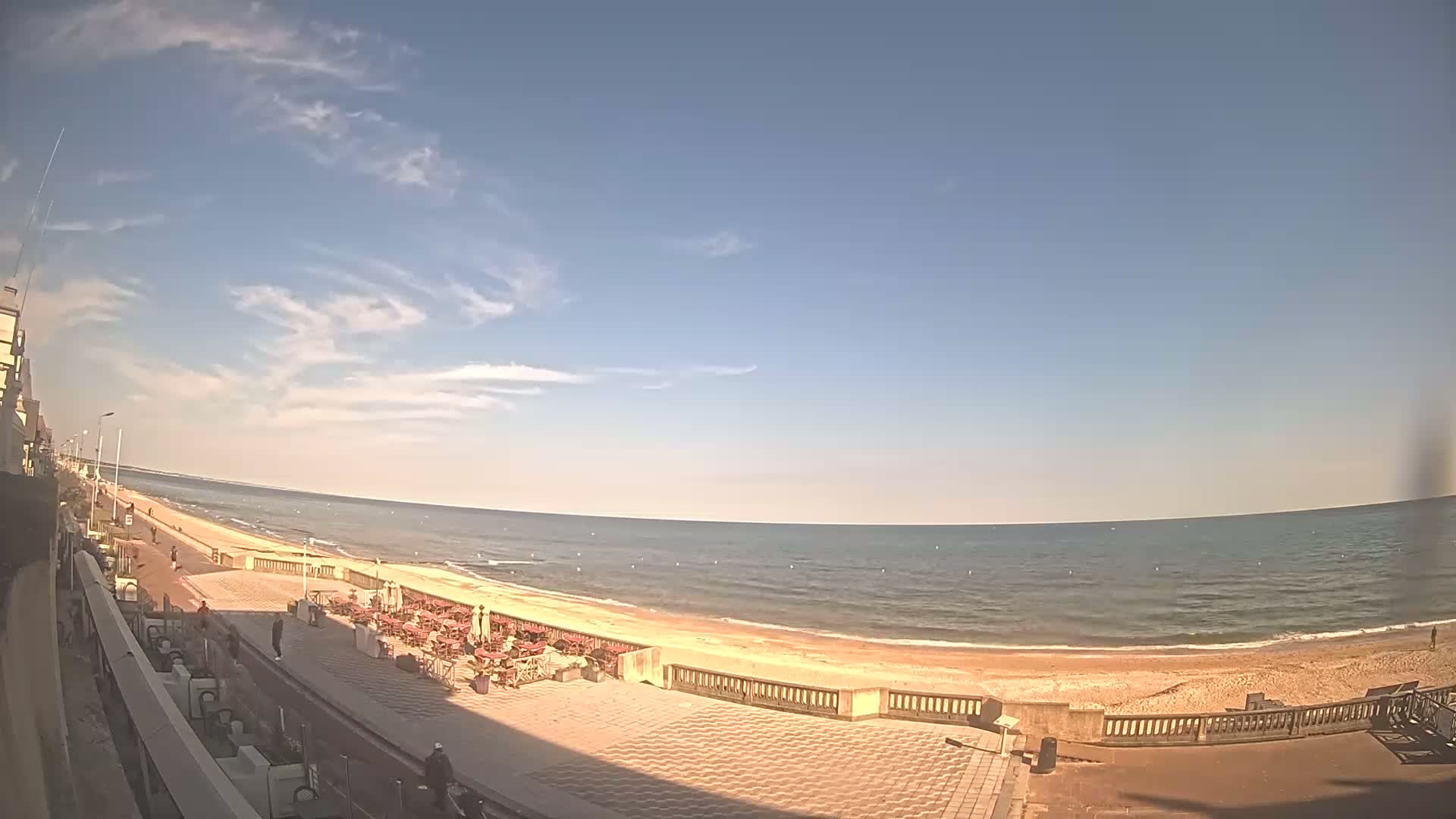 A mostly sunny day shows a wide sandy beach with calm ocean waves, a paved walkway lined with outdoor seating, and a few people strolling along the shore.