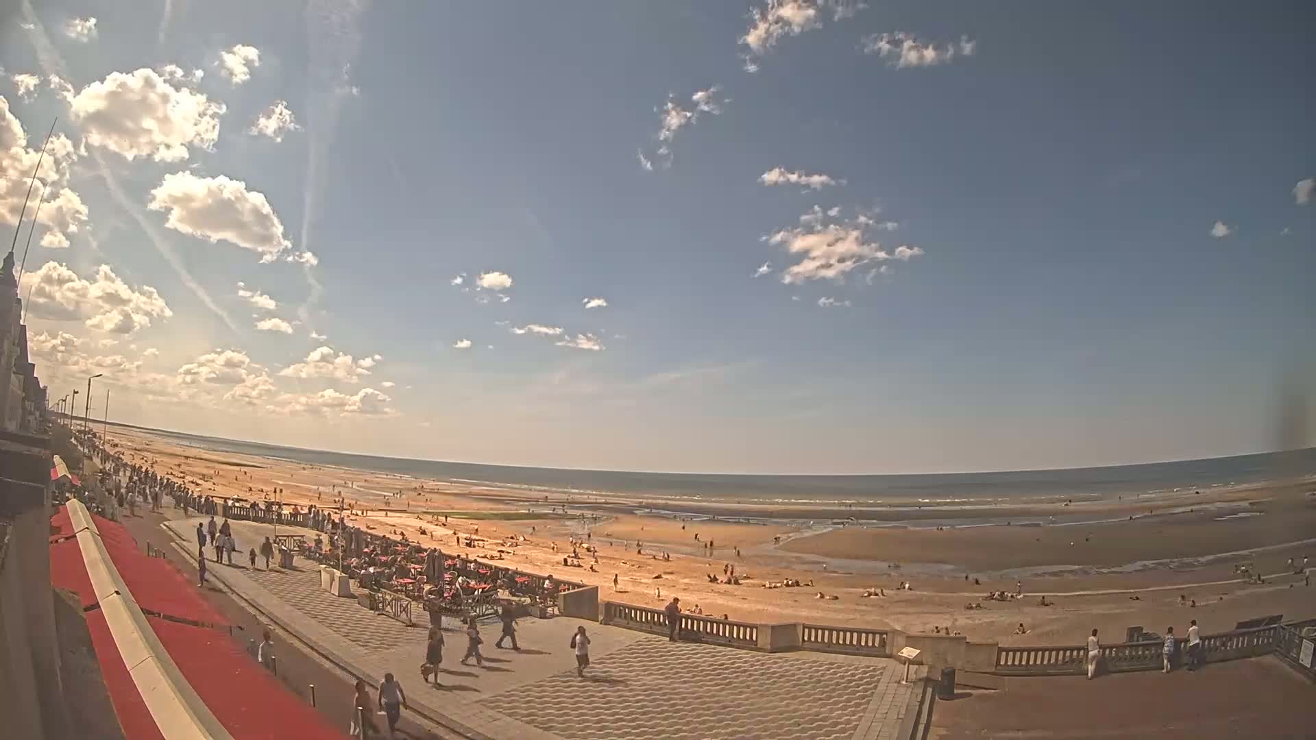 A sunny day at a beach shows many people on the sand and boardwalk under a partly cloudy sky.