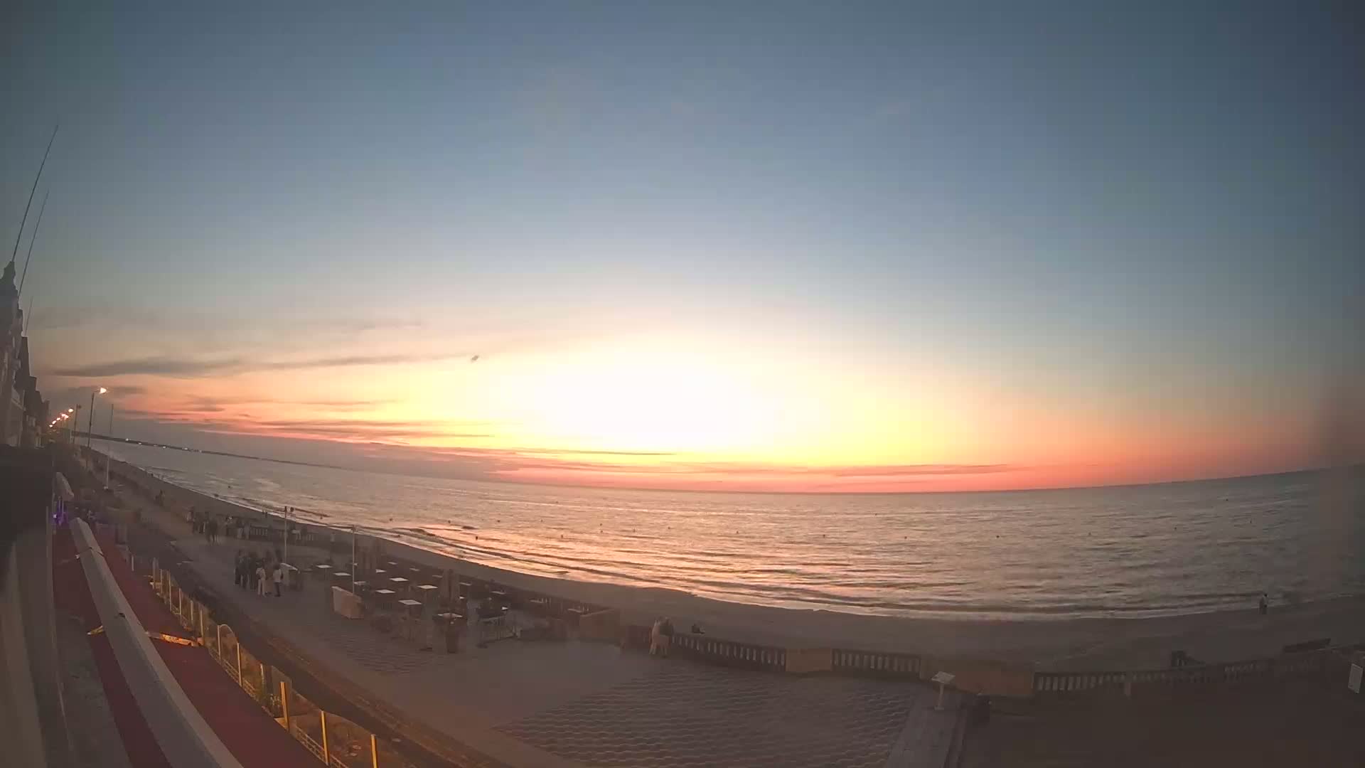 A beach scene at sunset shows a calm ocean with a sandy shoreline and a boardwalk with people walking, under a clear, mostly light-blue sky.