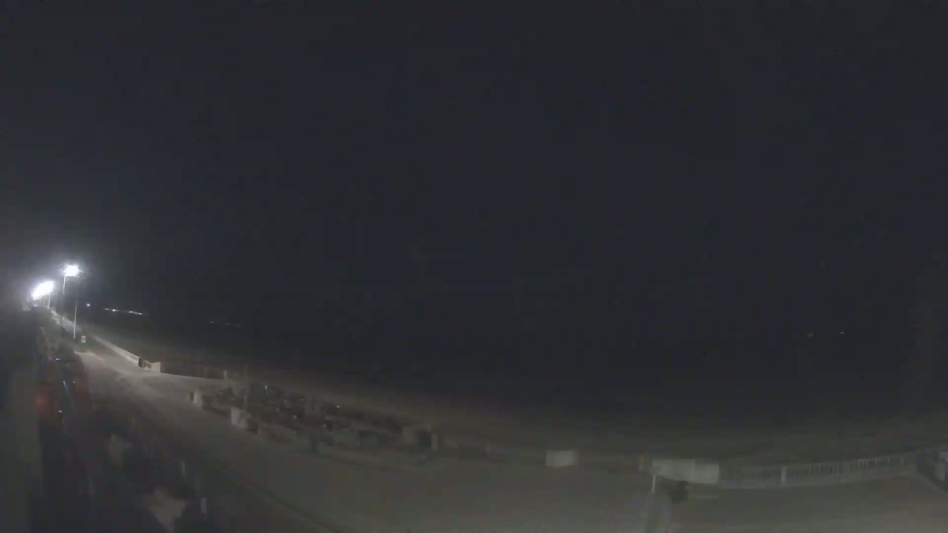 A nighttime view of a beach and boardwalk, illuminated by streetlights, under a dark, clear sky.