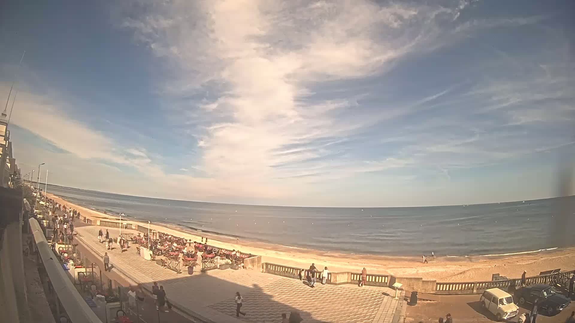A mostly sunny day at a beach with a promenade lined with outdoor seating and people, under a sky filled with wispy clouds.