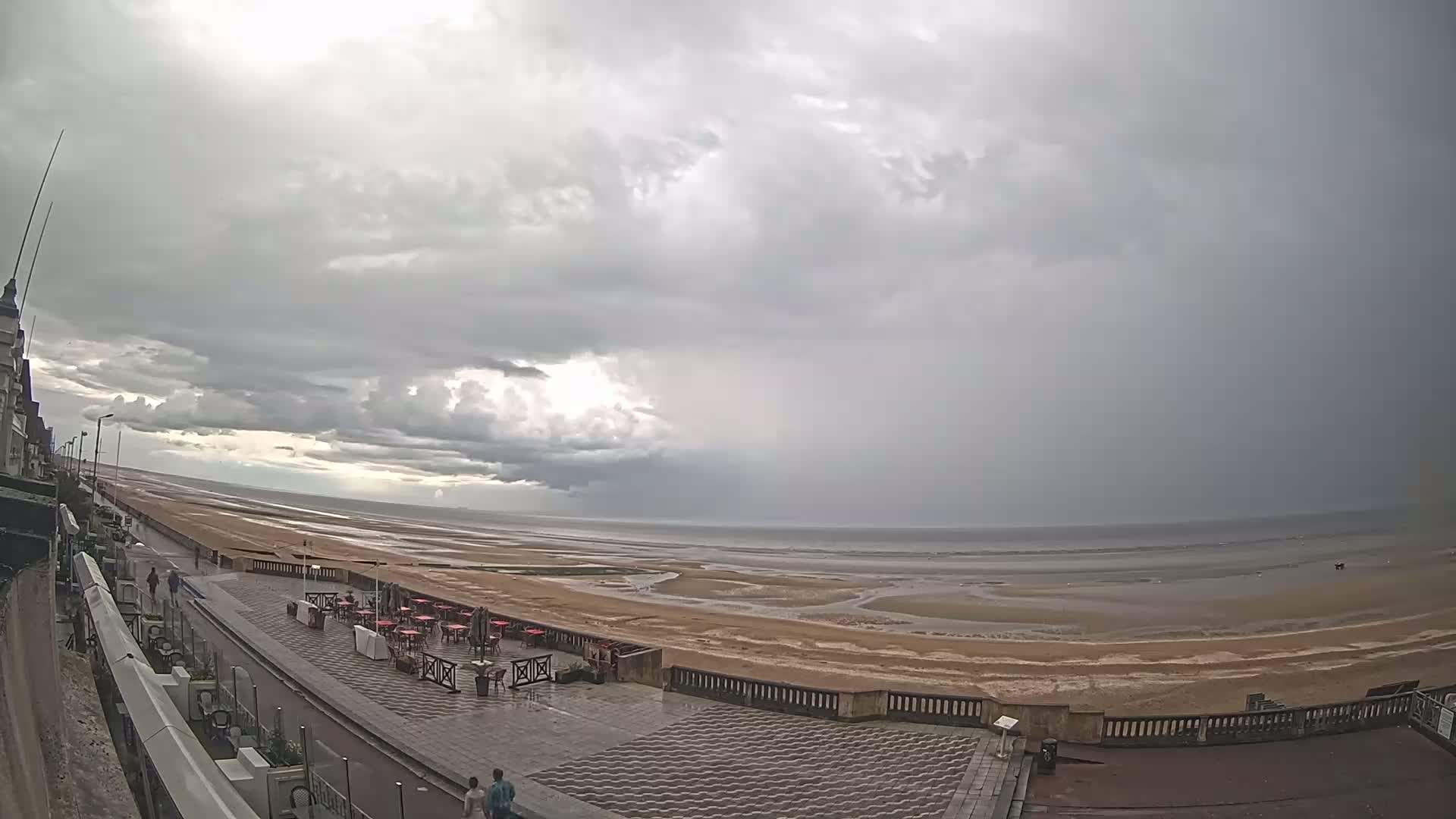A wide, overcast view of a beach with a promenade and outdoor seating area, showing a mostly flat, sandy beach exposed at low tide under a gray sky.