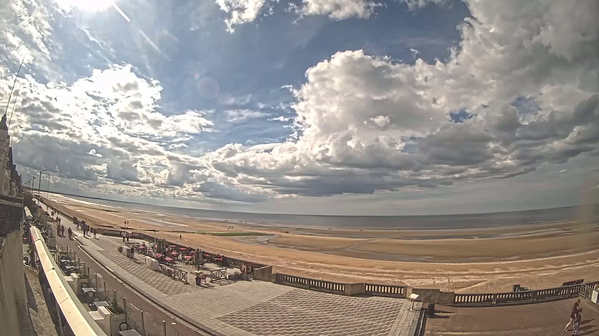A partly sunny day reveals a wide sandy beach and calm ocean, viewed from a seaside promenade with outdoor seating areas and people walking along the beachfront.