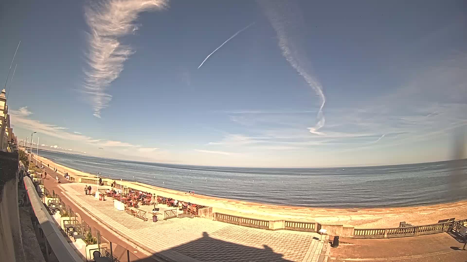 A mostly sunny day at a beachside promenade shows people relaxing at outdoor cafes next to a sandy beach and calm ocean under a mostly clear blue sky with wispy clouds and contrails.