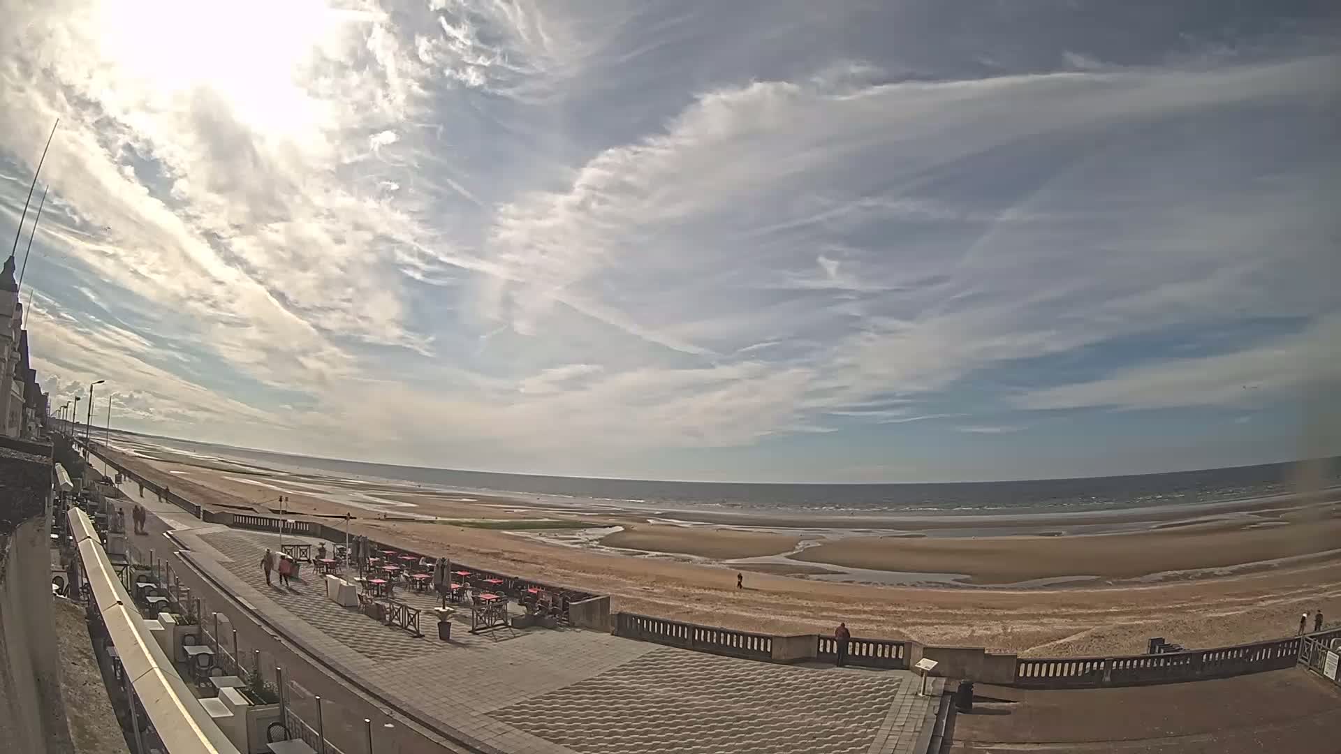 A wide, sunny shot shows a sandy beach and ocean under a mostly clear sky with thin, wispy clouds; an outdoor cafe sits along a paved walkway near the water.