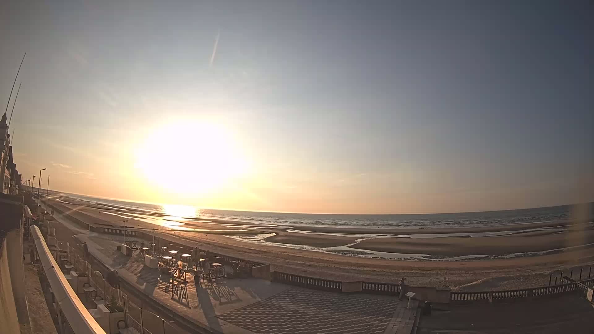 A wide shot of a beach at sunset, showing a sandy shore with small pools of water and an outdoor patio with tables and chairs near a building.