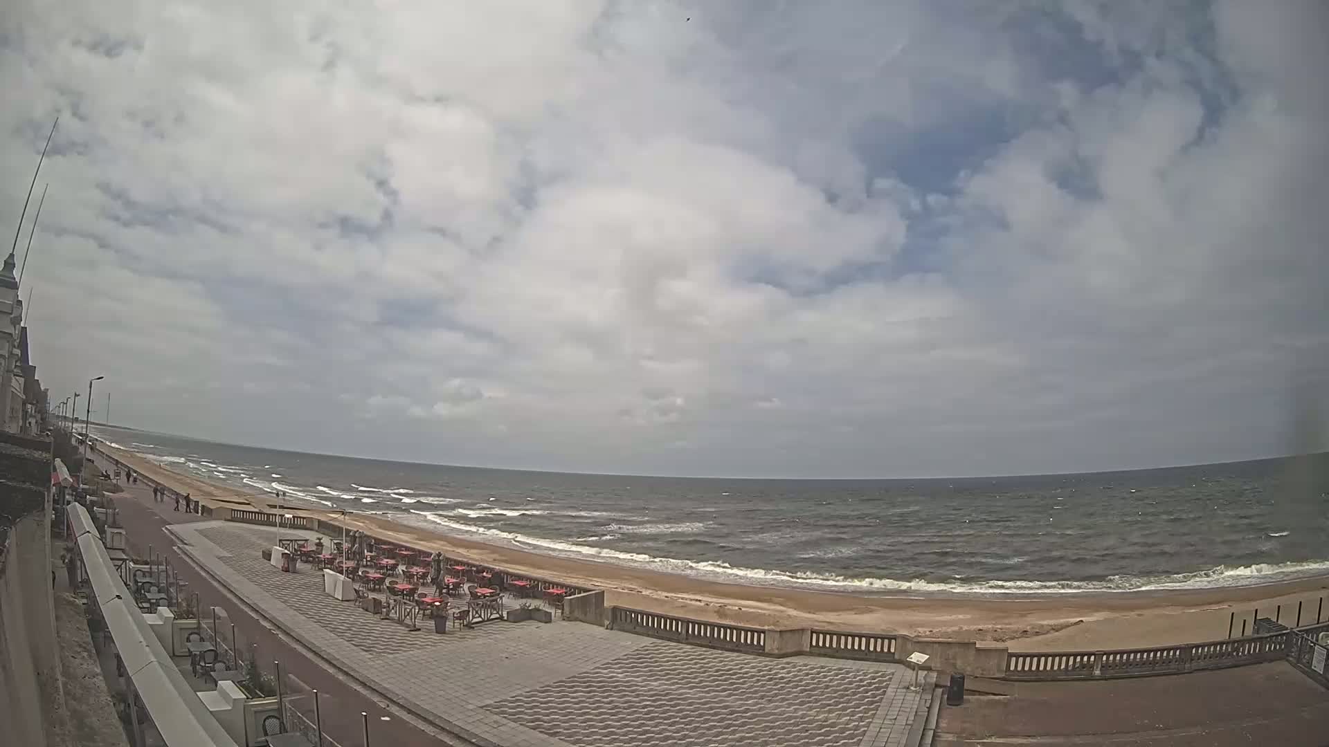A mostly cloudy sky overlooks a beach with moderate waves and outdoor seating areas along a paved walkway next to a building.