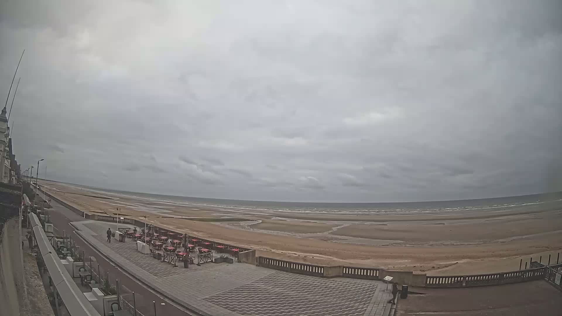 A wide, overcast view shows a sandy beach extending to a calm sea, with an outdoor cafe and promenade nearby.