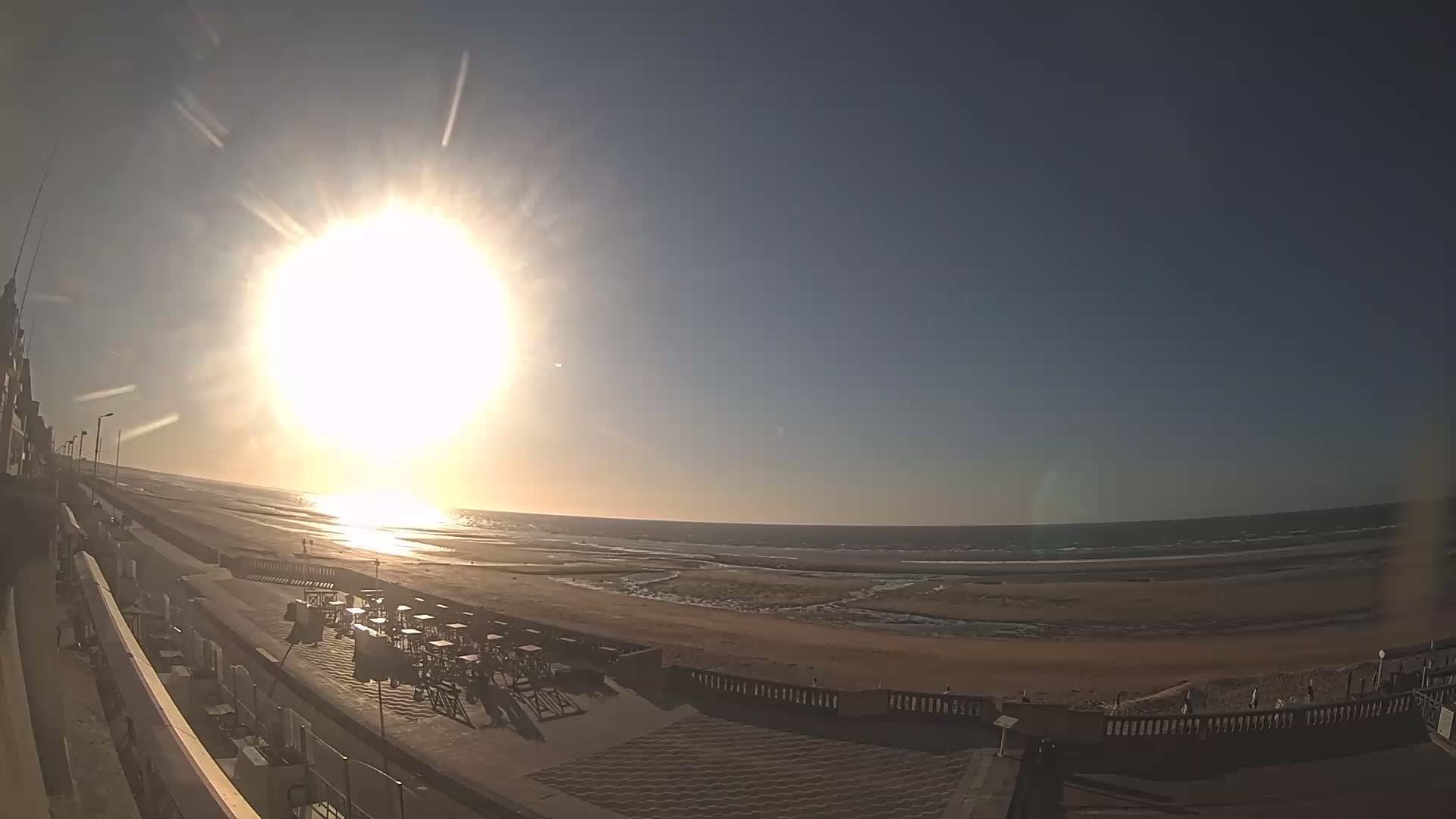 A bright sun shines over a beach with an outdoor patio area containing empty tables and chairs.