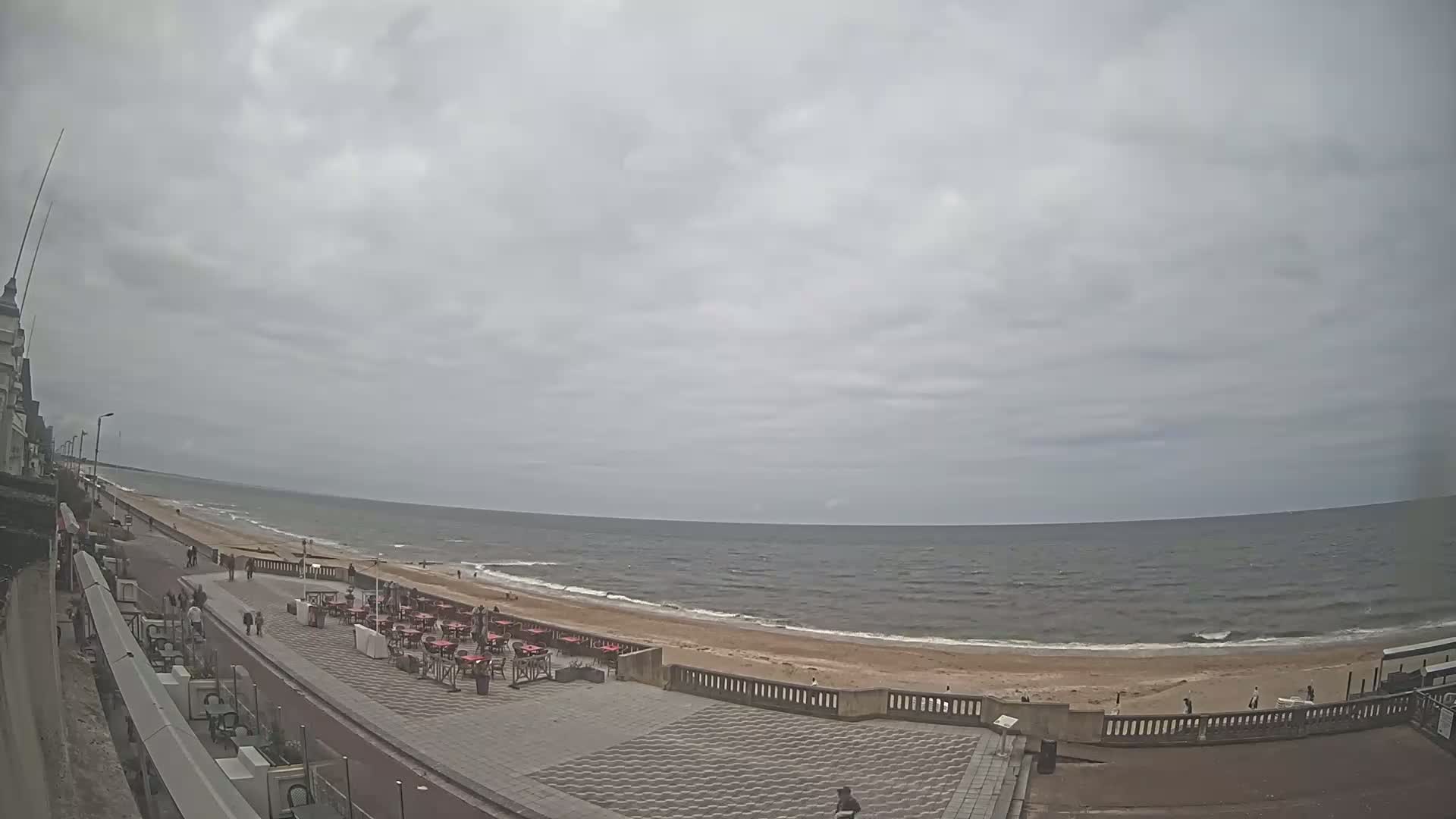 A paved beachfront promenade with outdoor seating overlooks a sandy beach and a somewhat choppy sea under a cloudy sky.