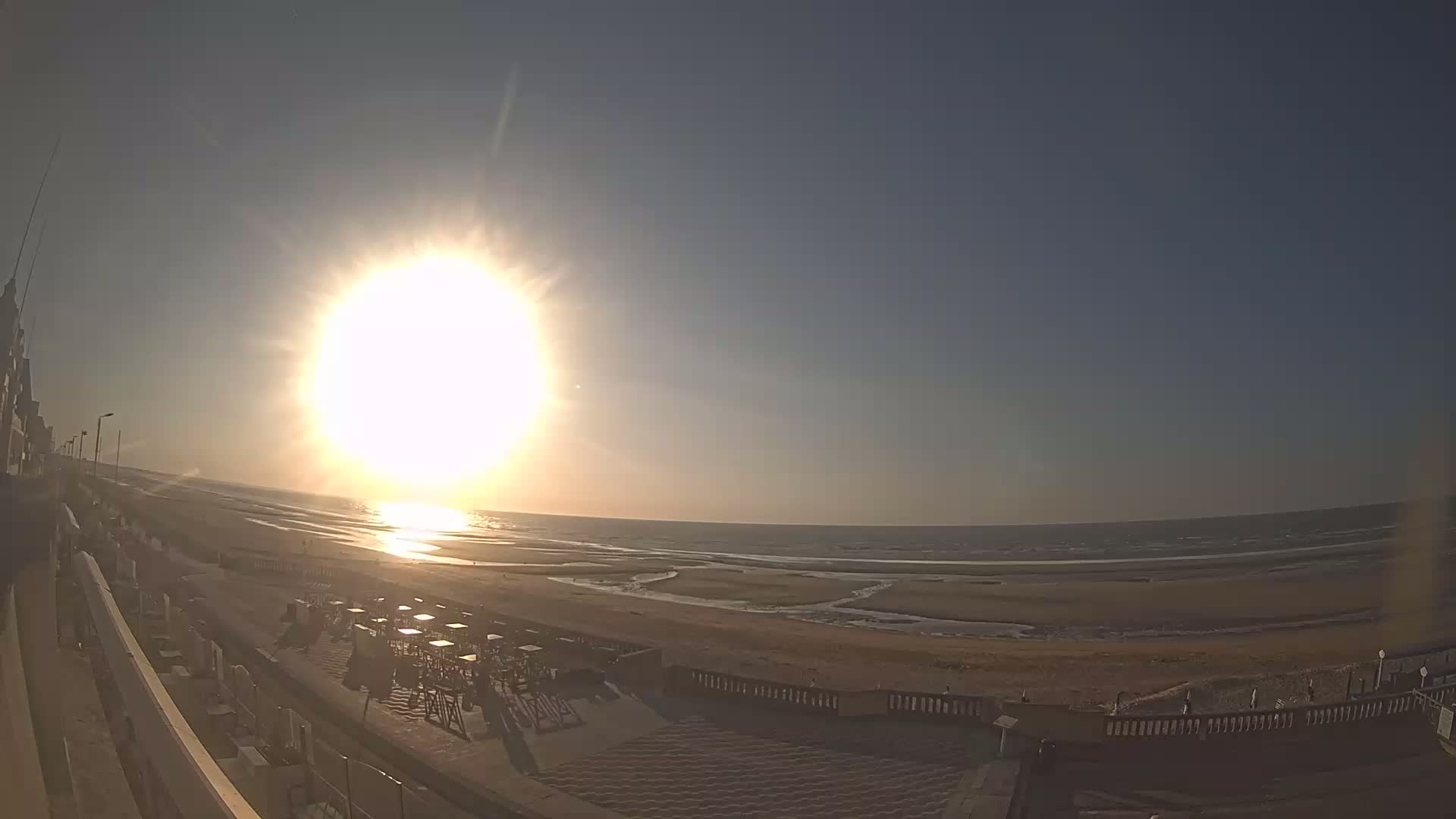 A bright sun shines over a beach with a mostly calm ocean, and outdoor seating near a building.
