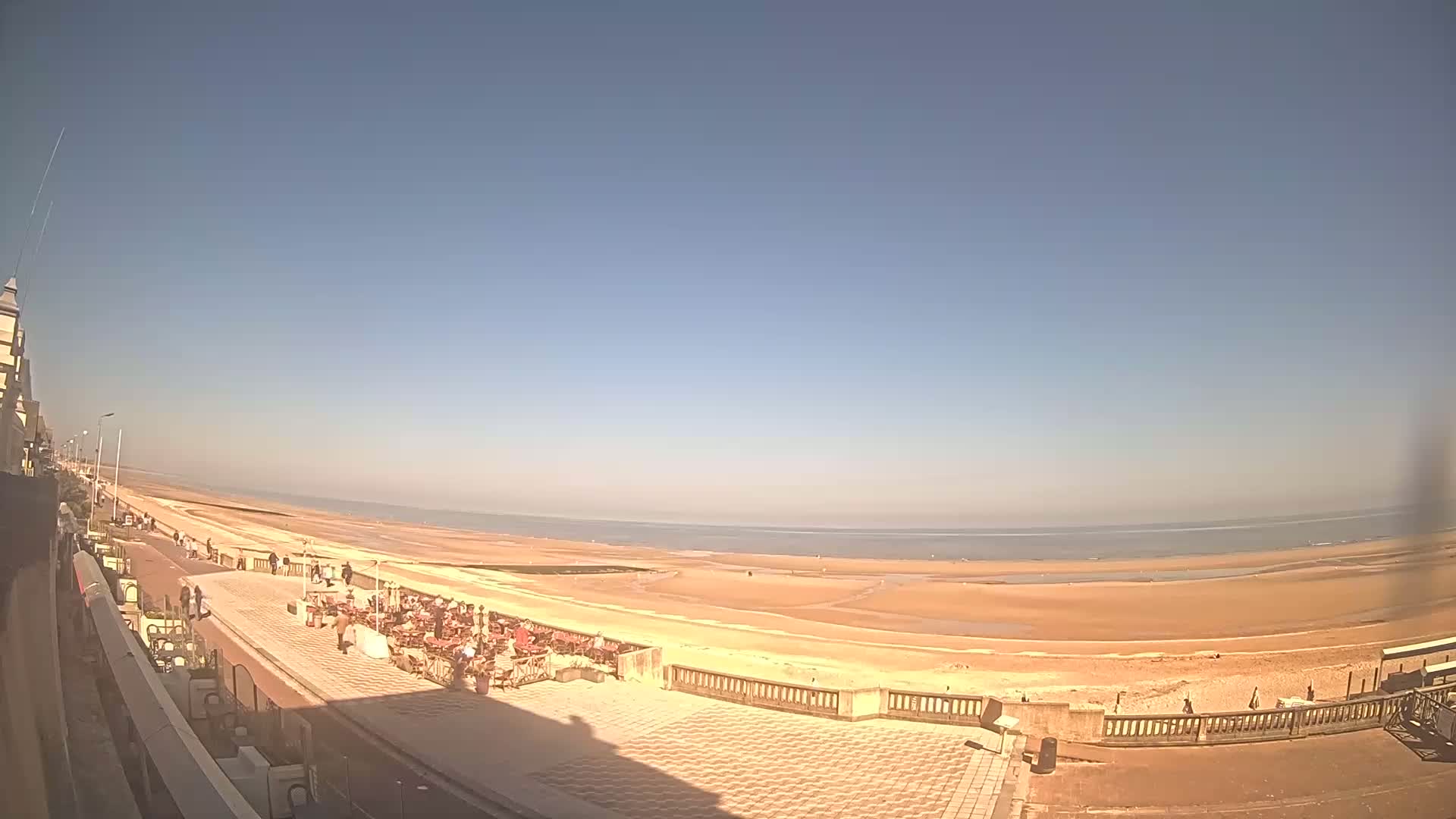 A wide, sandy beach stretches along a calm ocean under a clear, bright blue sky, with people strolling along a paved boardwalk lined with outdoor seating.