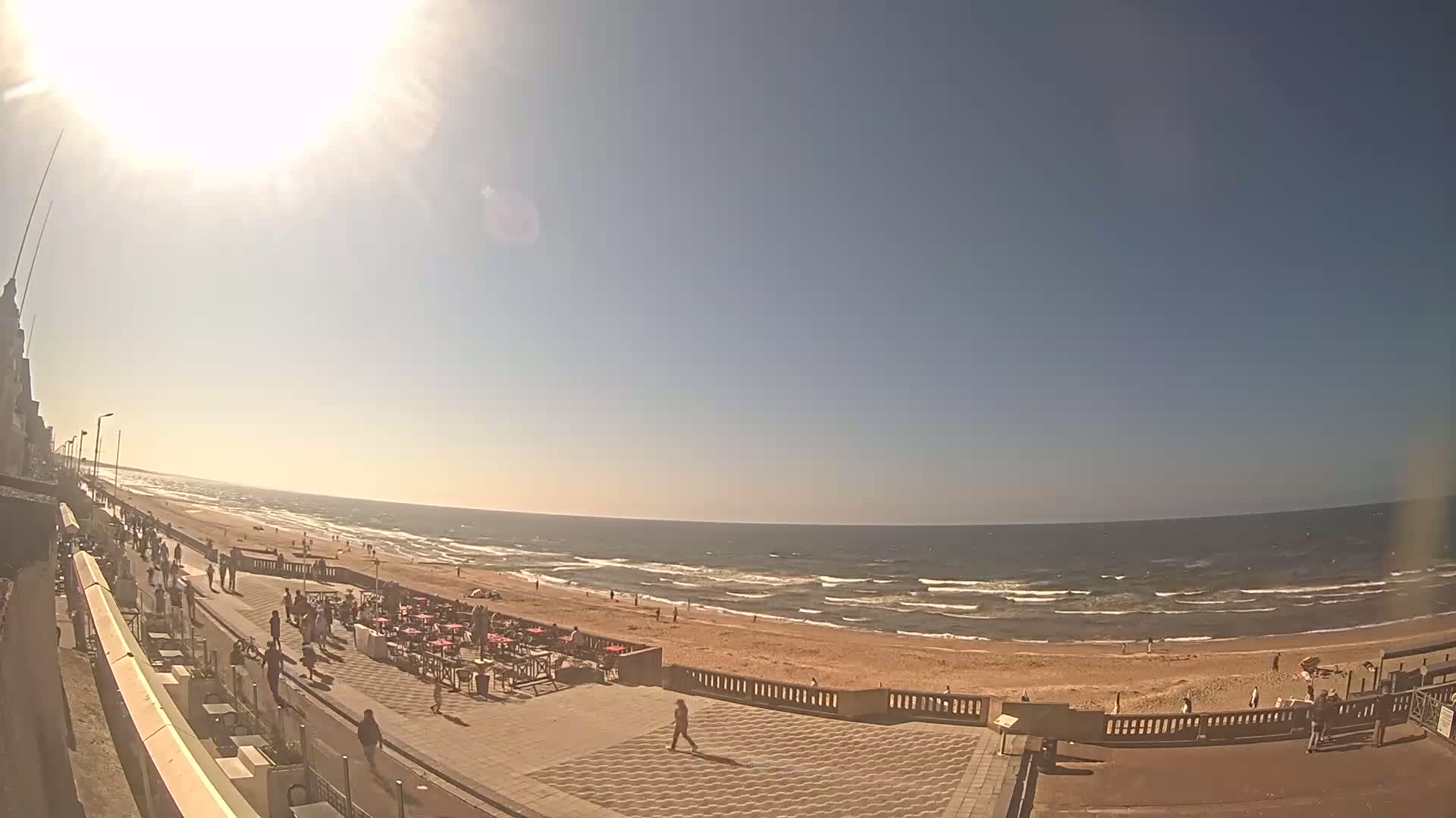 A sunny day at a beach shows a sandy shore with small waves, people walking along a paved promenade near outdoor seating, and a building in the background.