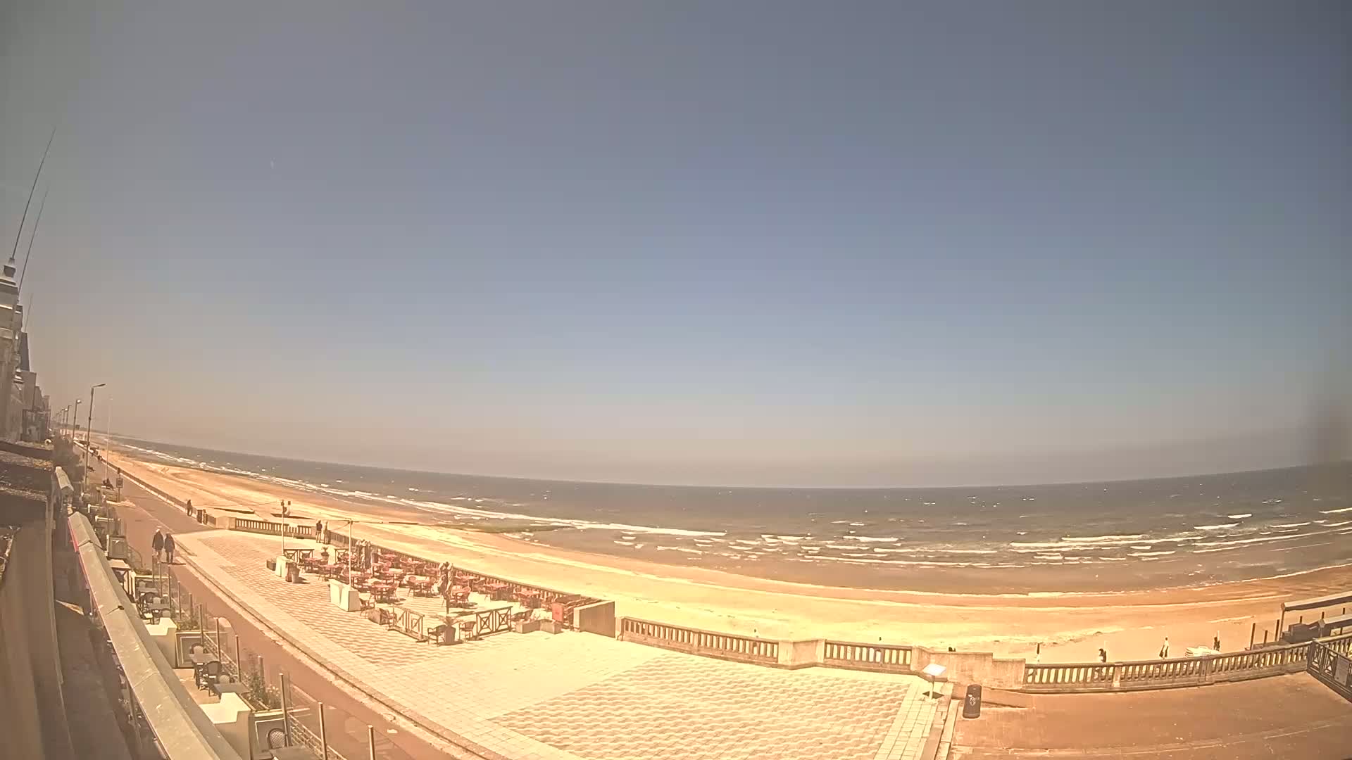 A sandy beach with a calm sea under a clear, sunny sky is seen from a high vantage point, overlooking a paved promenade with outdoor seating.